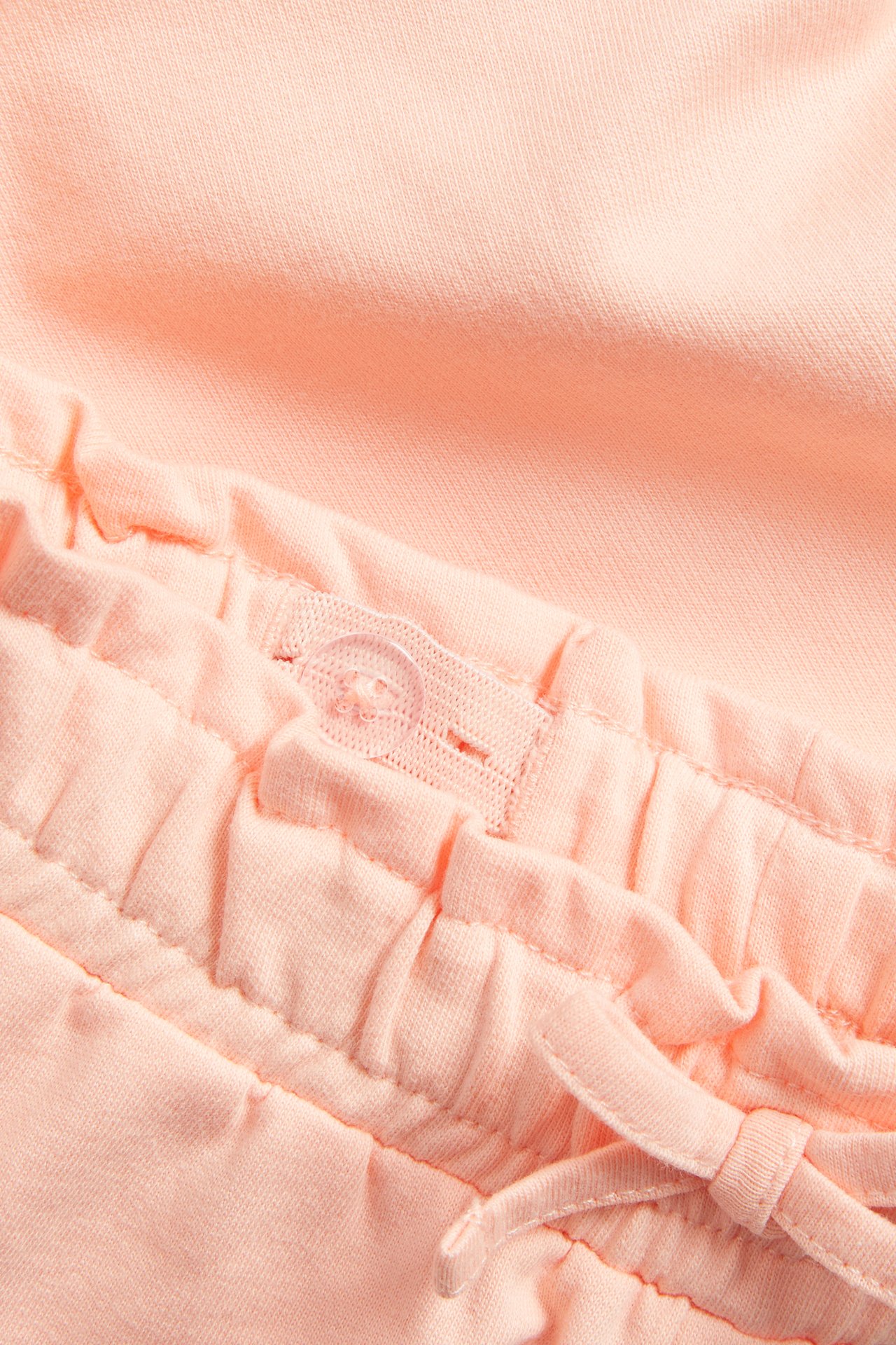 Nærbilde av korallfargede bomullsshorts med elastisk linning, justerbar knapp og dekorativ knyting.