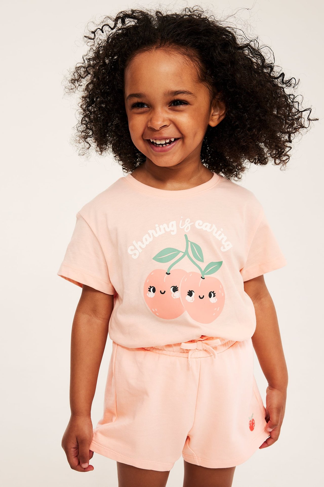 Widok z przodu uśmiechniętego dziecka w jasnych koralowych szortach z haftem truskawki i pasującej koszulce w brzoskwinie.