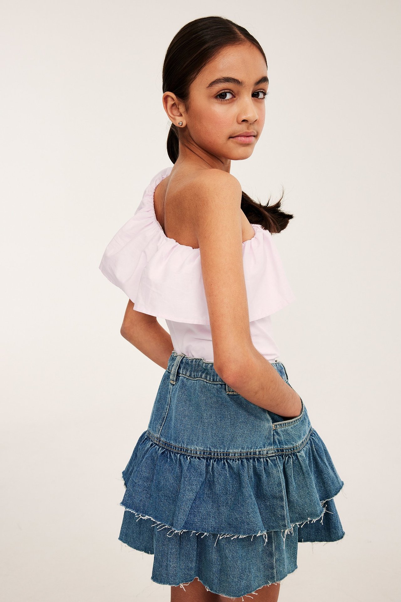 En flicka bär en ljusrosa volangtopp med en axel i bomull och en blå jeanskjol med volanger. Sidovy.