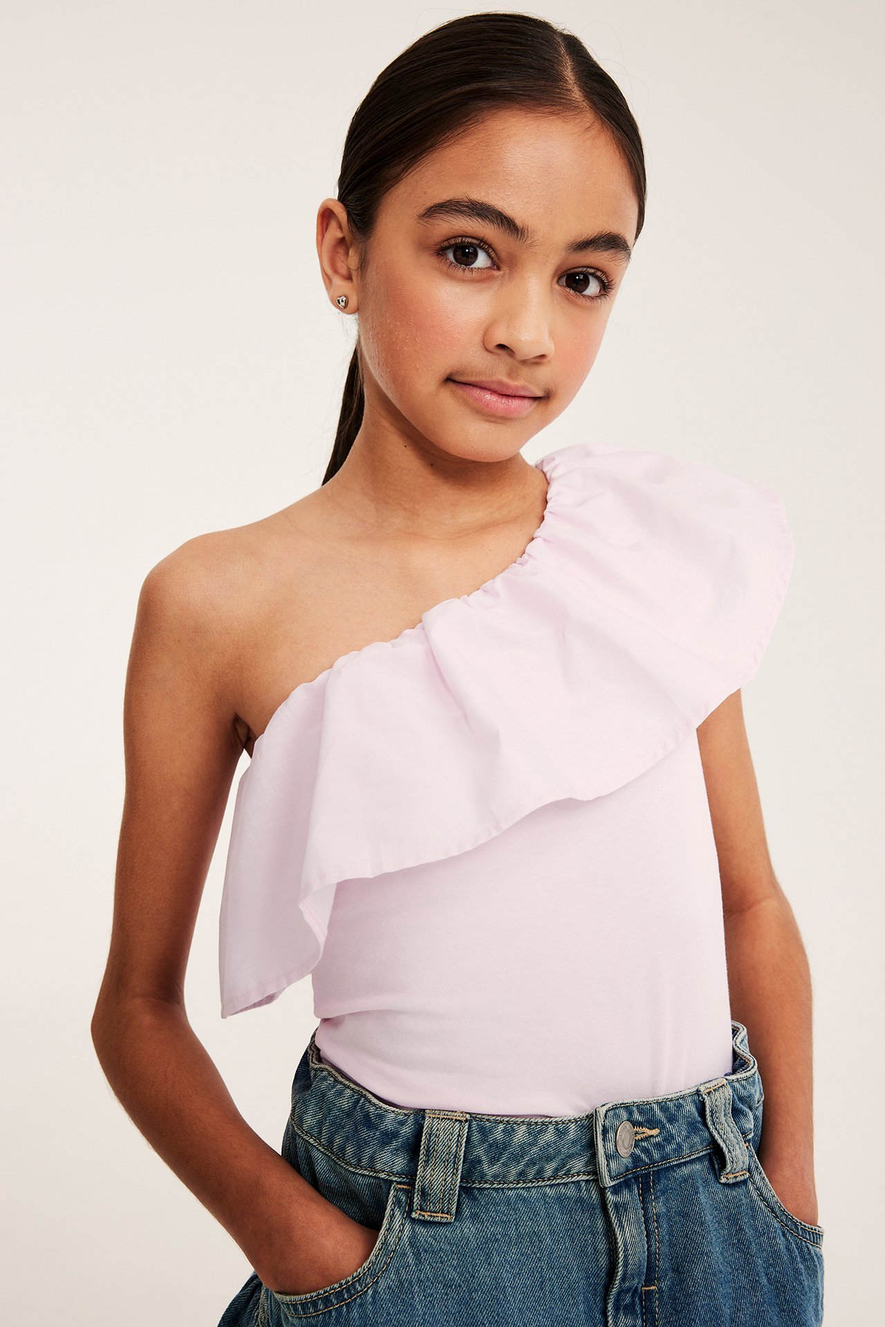 Frontvy av ett barn i ljusrosa volangtopp i bomull med en axel och blå jeans.