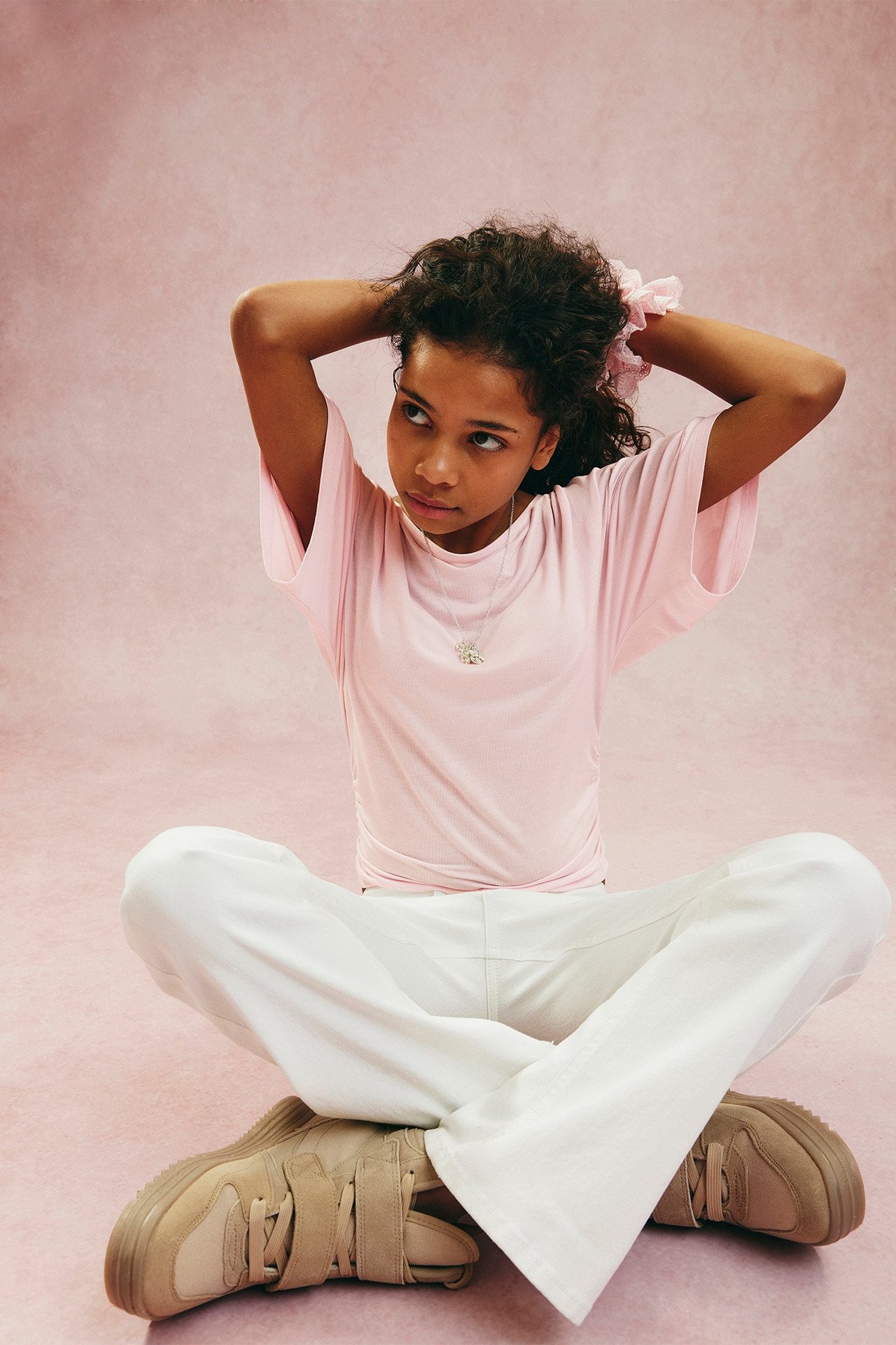 Przód dziecka w jasnoróżowym drapowanym topie z rękawami motylkowymi, białych spodniach i beżowych sneakersach.