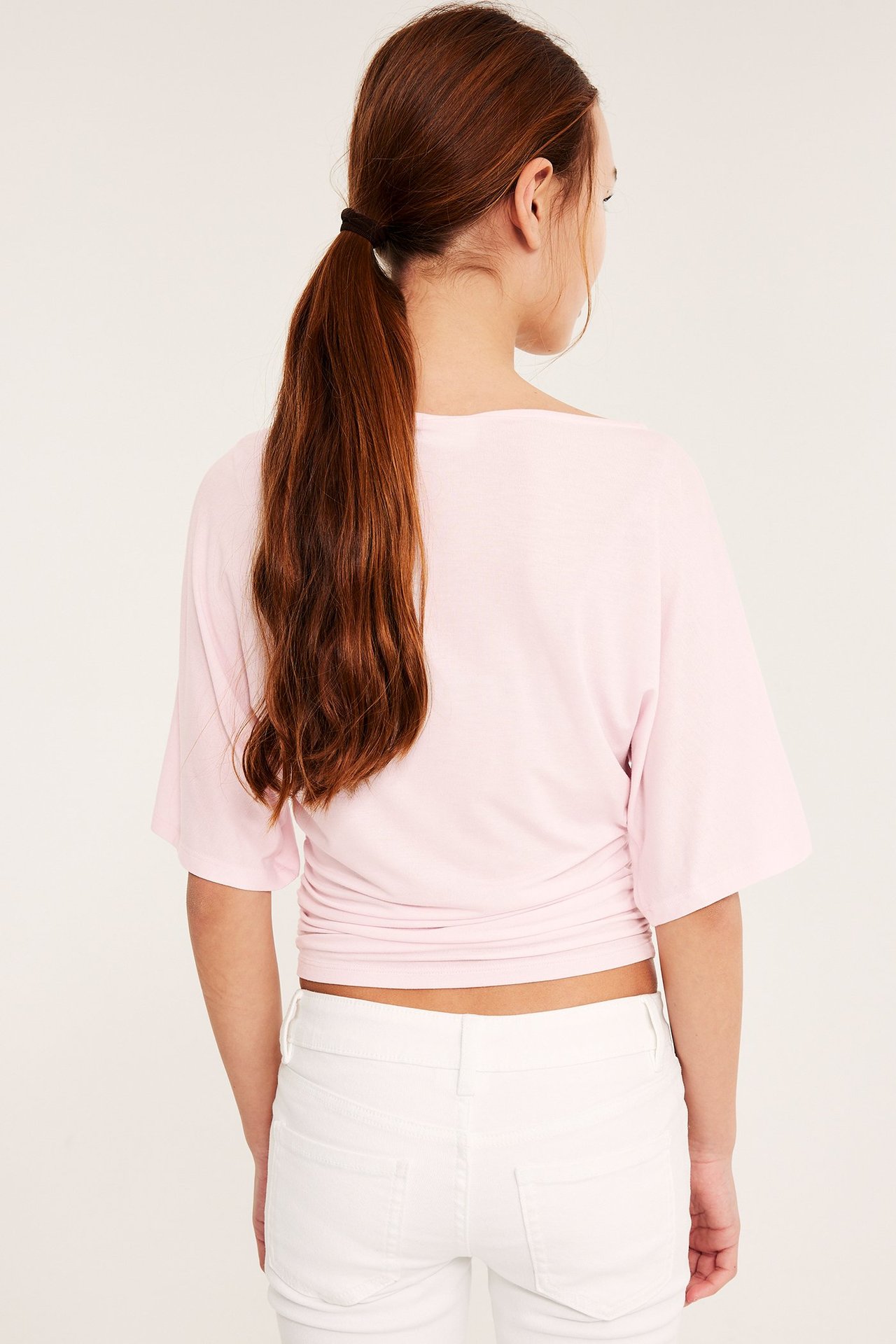 Ryggvy: Barn i ljusrosa draperad topp med fjärilsärmar, rynkad midja och vita jeans.