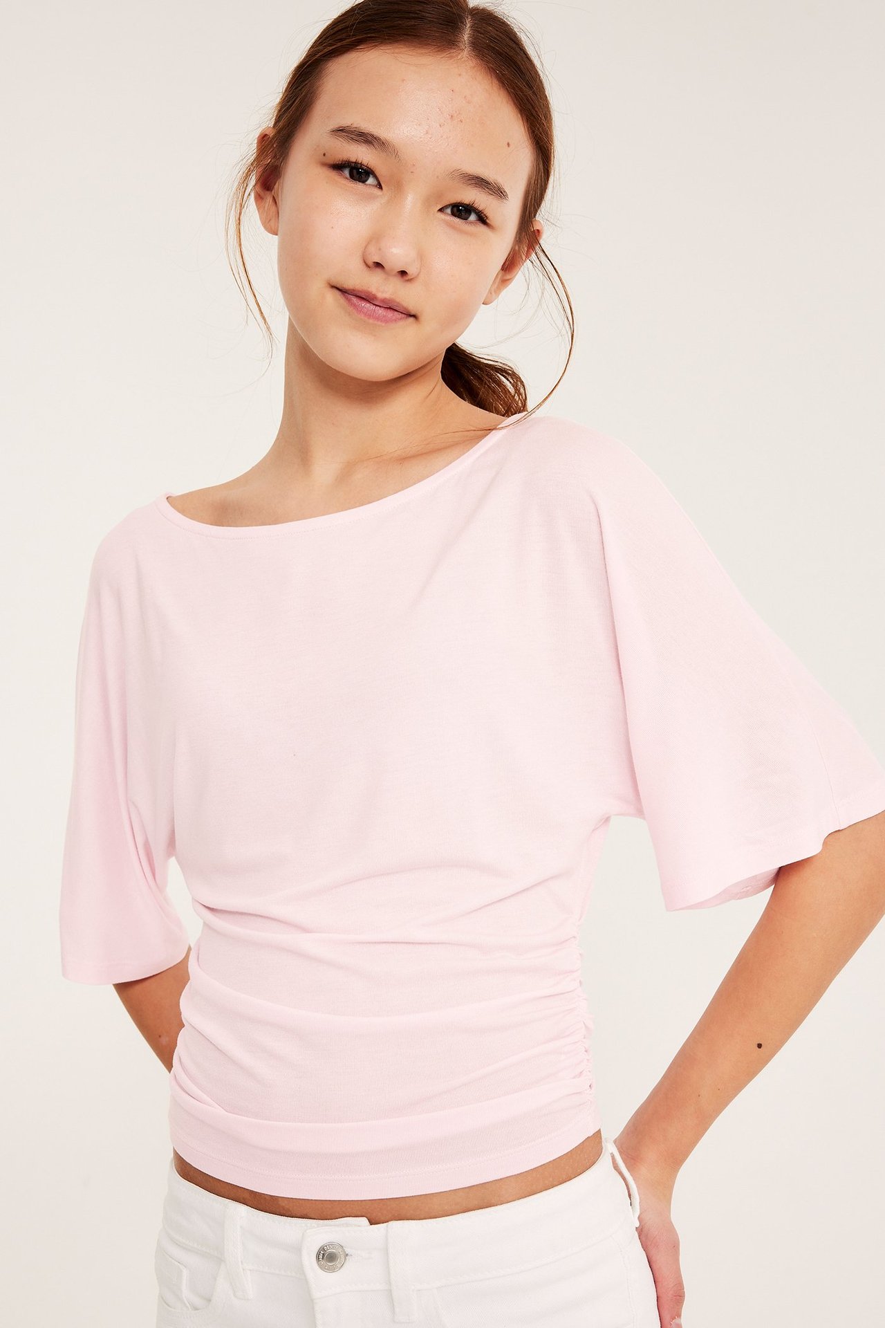 Framifrån: Ett barn bär en ljusrosa draperad topp med fjärilsärmar, rynkad midja, och vita jeans.