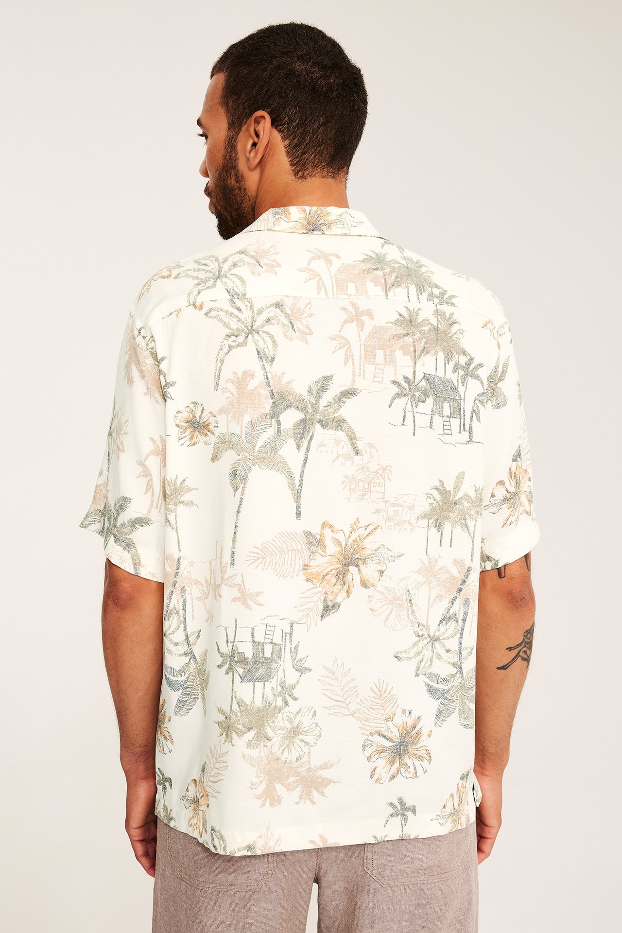 Ryggvy av man i ljusbeige, tropiskt mönstrad kortärmad resortskjorta och bruna shorts.