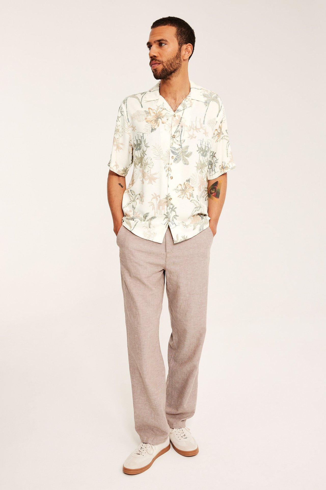 Framifrån: en man i en ljusbeige mönstrad kortärmad resortsjorta, ljusbruna byxor och beiga sneakers.