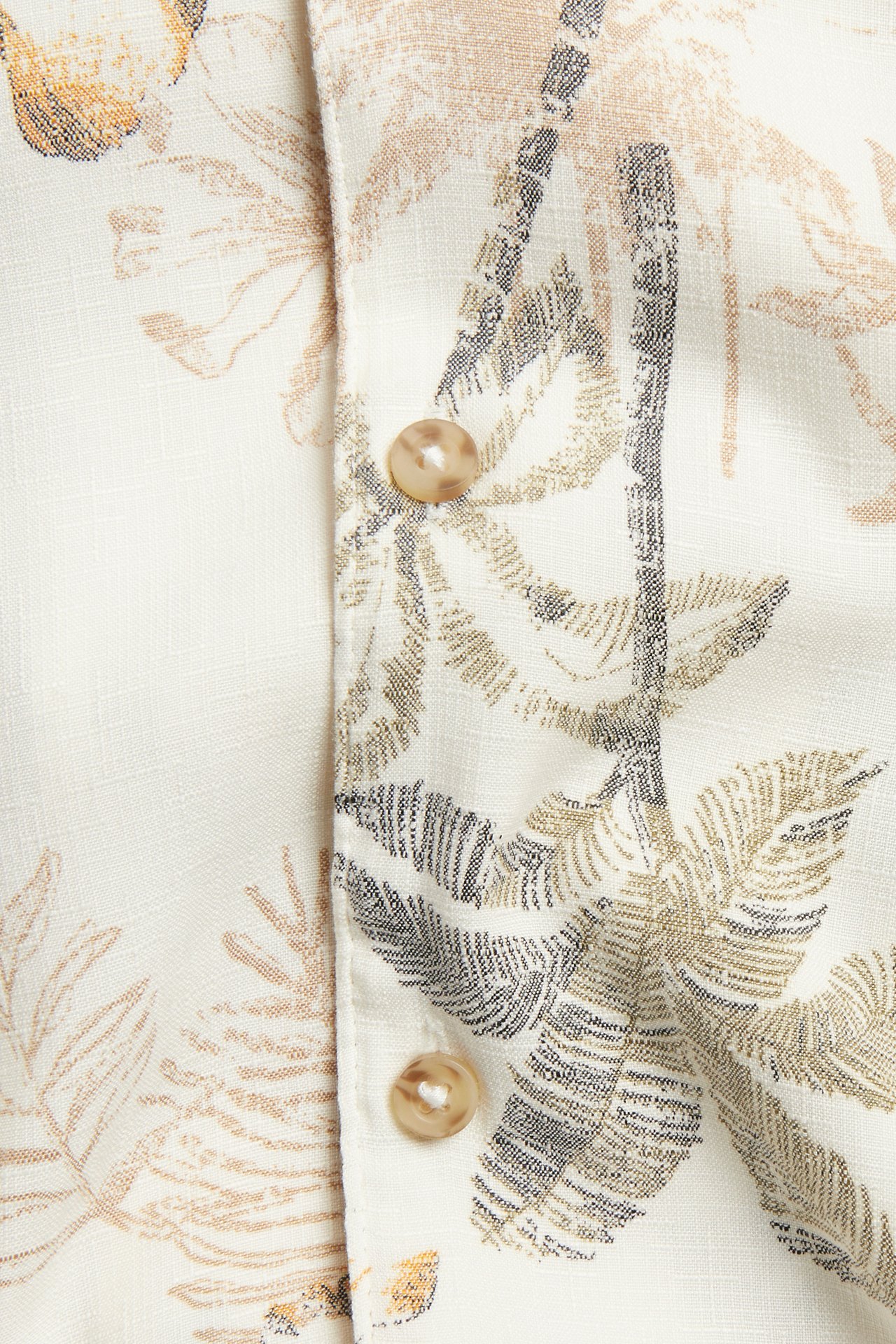 Lähikuva vaaleanbeigestä lyhythihaisesta resort-paidasta, jossa on ruskeat napit ja palmukuvio.