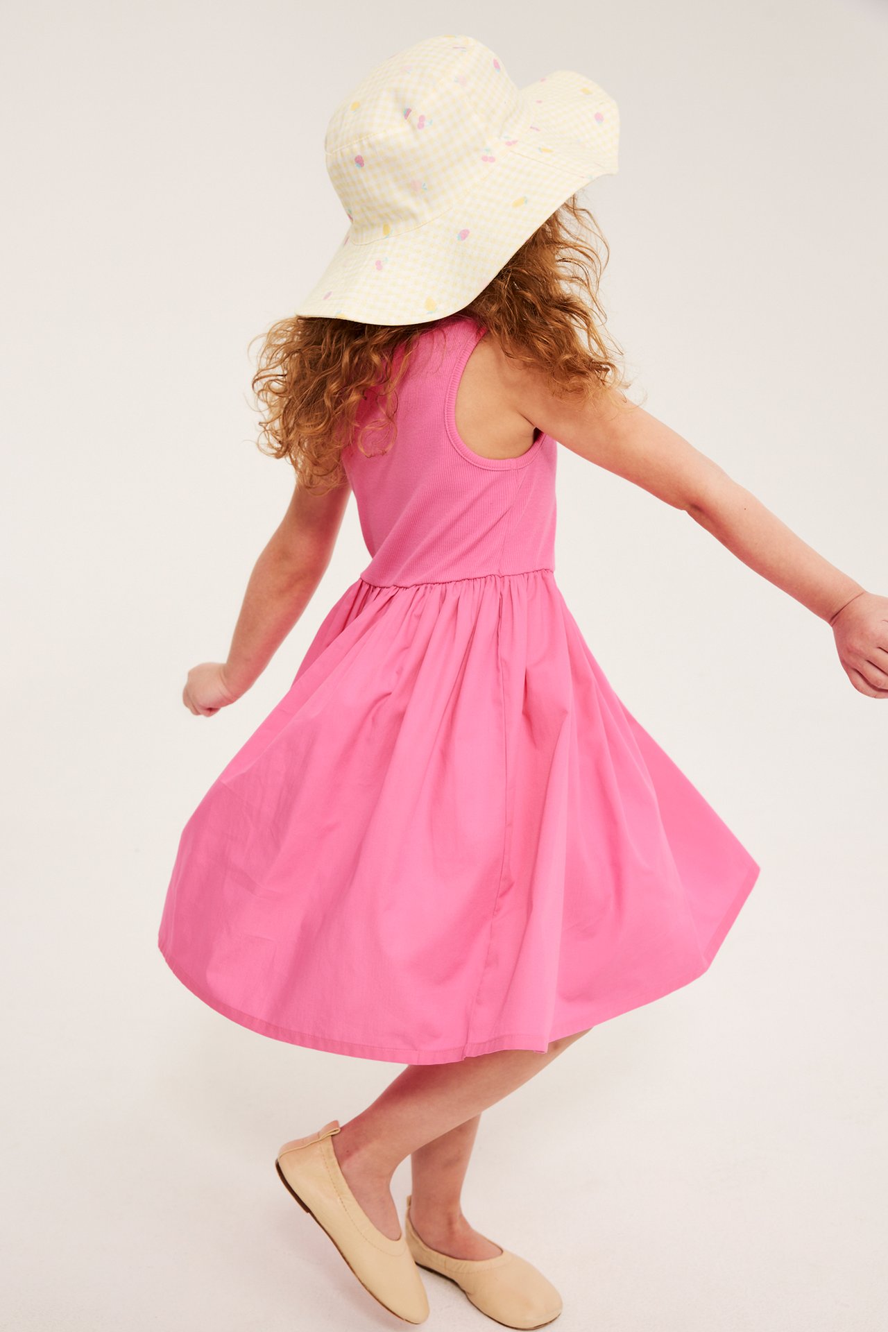 Widok z tyłu: Dziecko w różowej bawełnianej sukience bez rękawów, wzorzystym kapeluszu i beżowych baletkach.