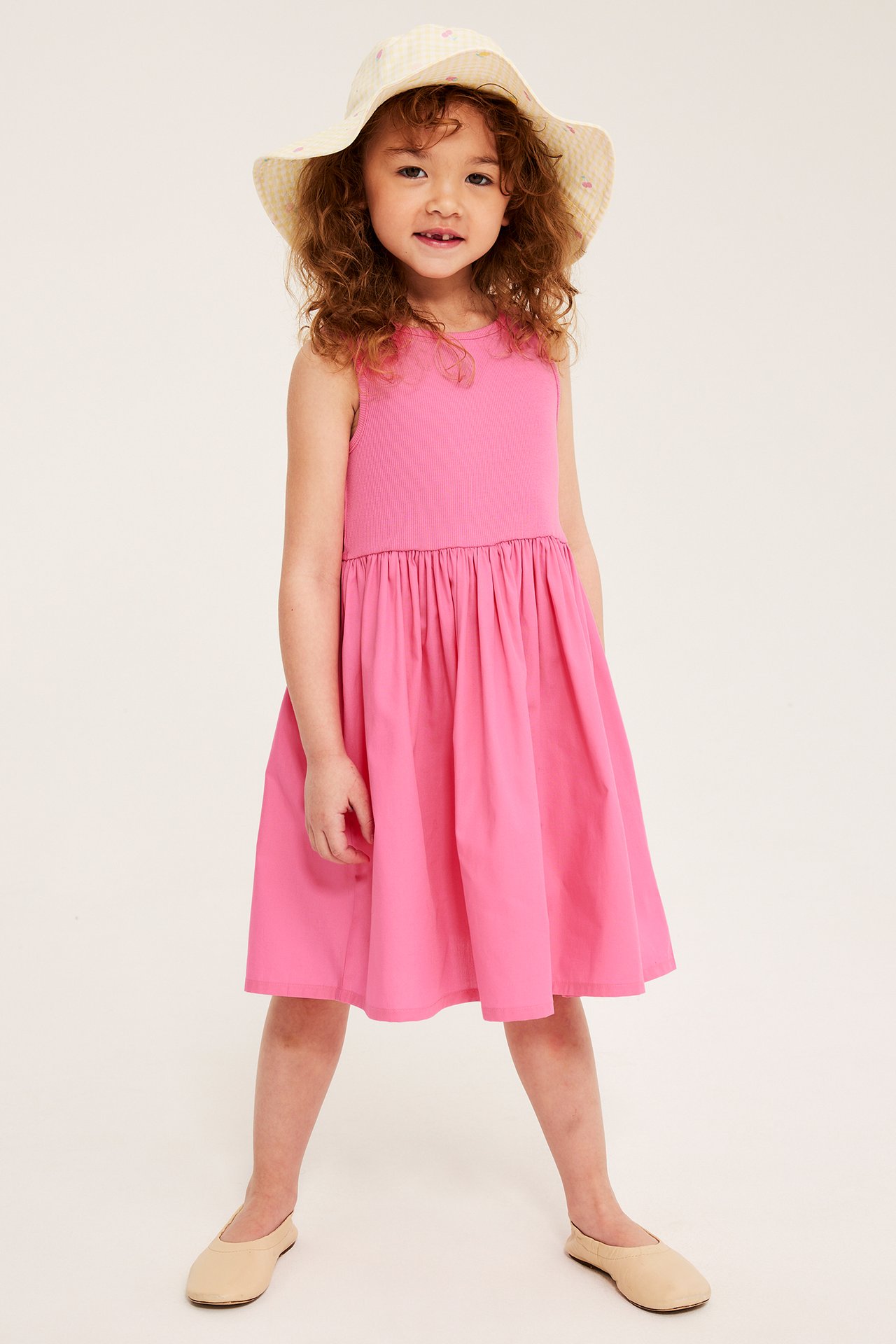 Framifrån: Barn bär rosa ärmlös bomullsklänning, gulrutig solhatt och beige ballerinaskor.
