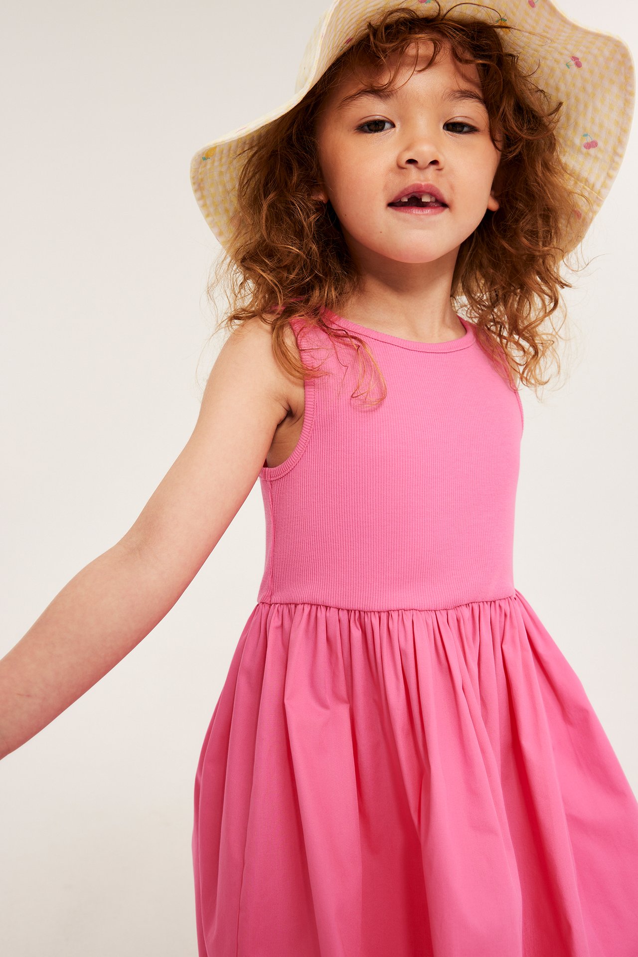 Zbliżenie na dziecko w różowej, bawełnianej sukience bez rękawów i żółtym kapeluszu z wiśniami.