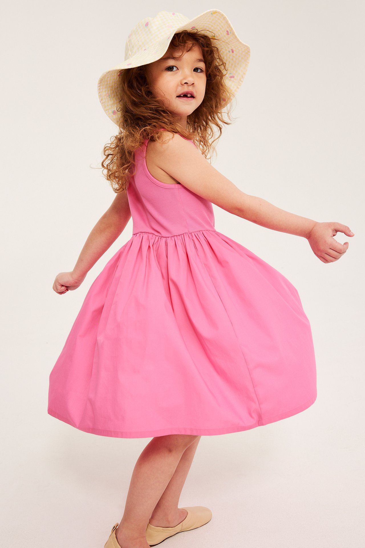Barn i framifrån i en rosa ärmlös bomullsklänning, en gulrutig solhatt och beiga platta skor.