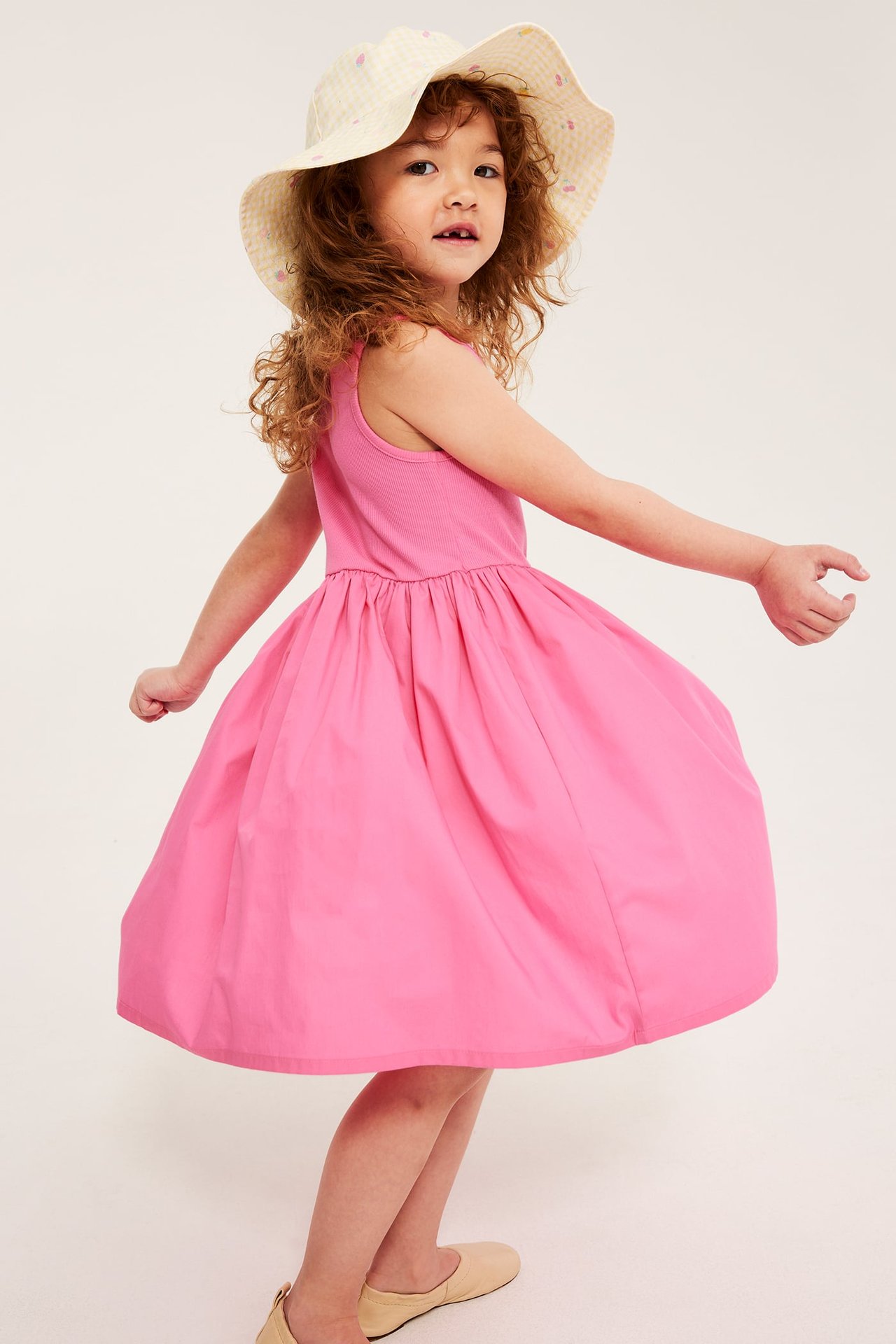Dziecko przodem w różowej bawełnianej sukience bez rękawów, żółtym kapeluszu i beżowych płaskich butach.
