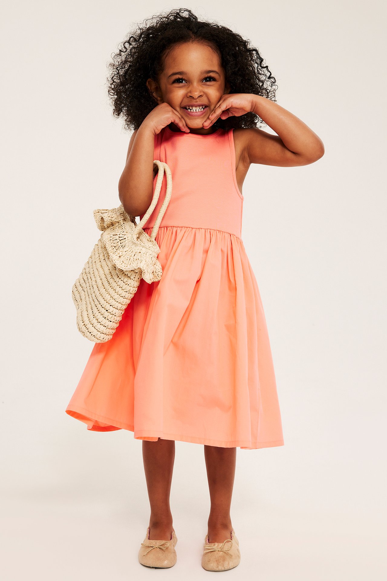Framifrån, ett leende barn i en ljusorange ärmlös klänning, håller beige stråväska och ljusbruna ballerinaskor.
