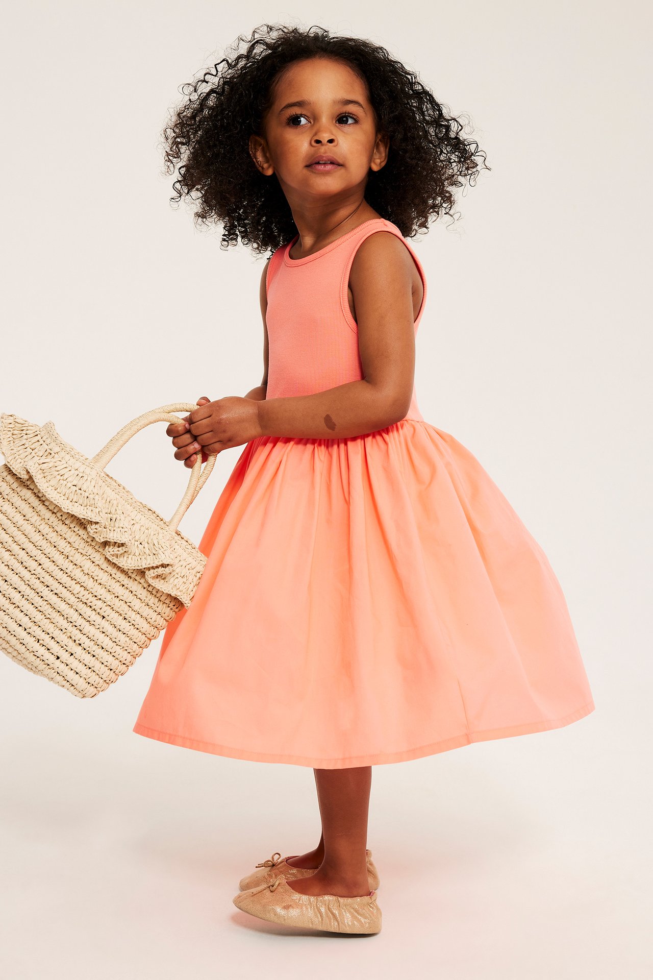 Framsida på ett barn i en ljusorange bomullsklänning, håller en stråväska och med guldiga ballerinaskor.