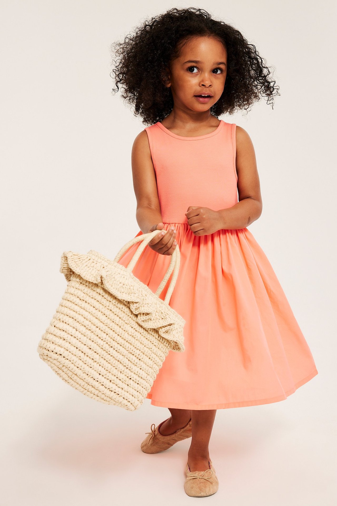 Dziecko w jasnopomarańczowej sukience bez rękawów, z beżową słomianą torbą, w brązowych balerinach.