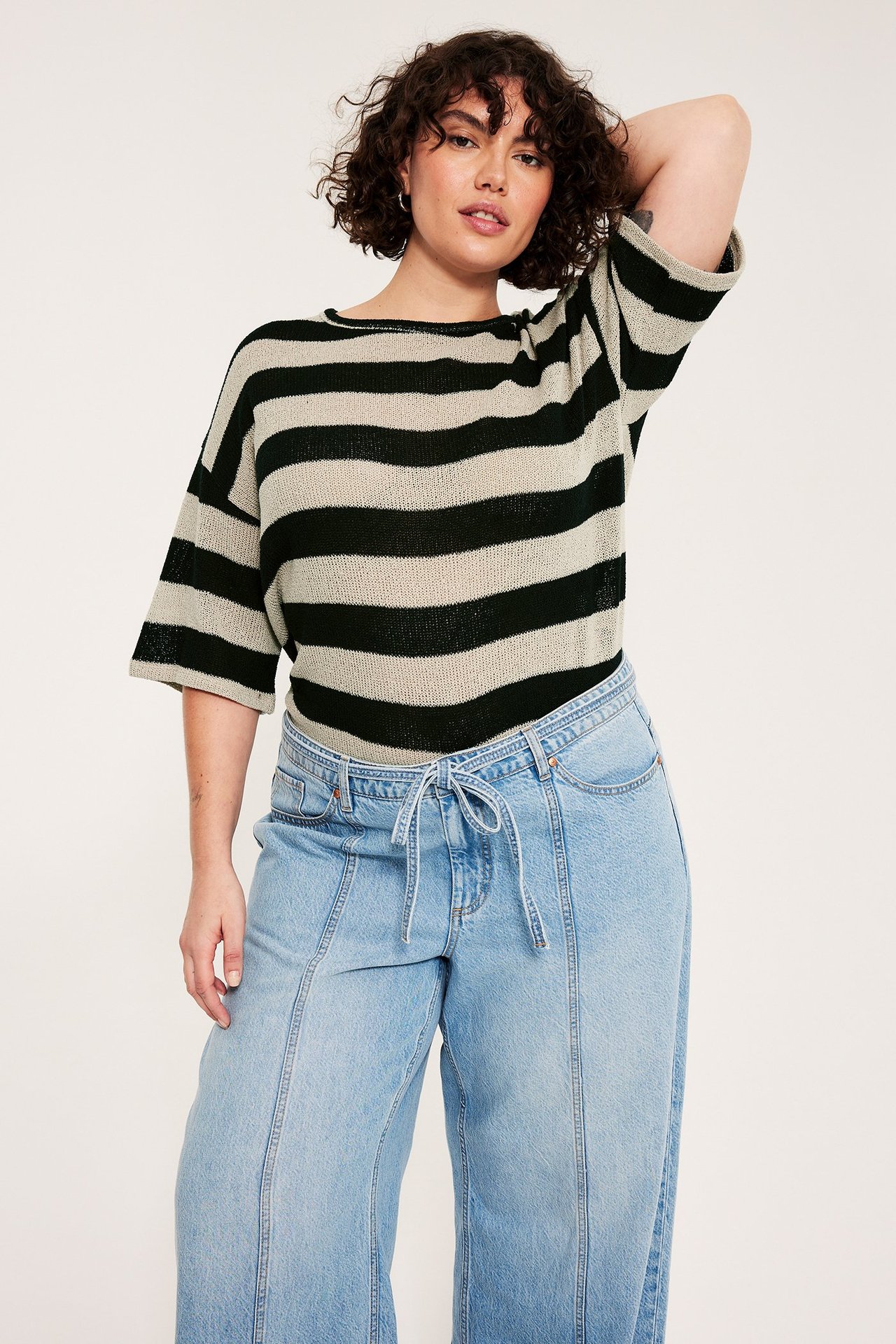 Forfra, kvinne i svart- og lys beige stripet strikket kortermet topp og lyseblå jeans.