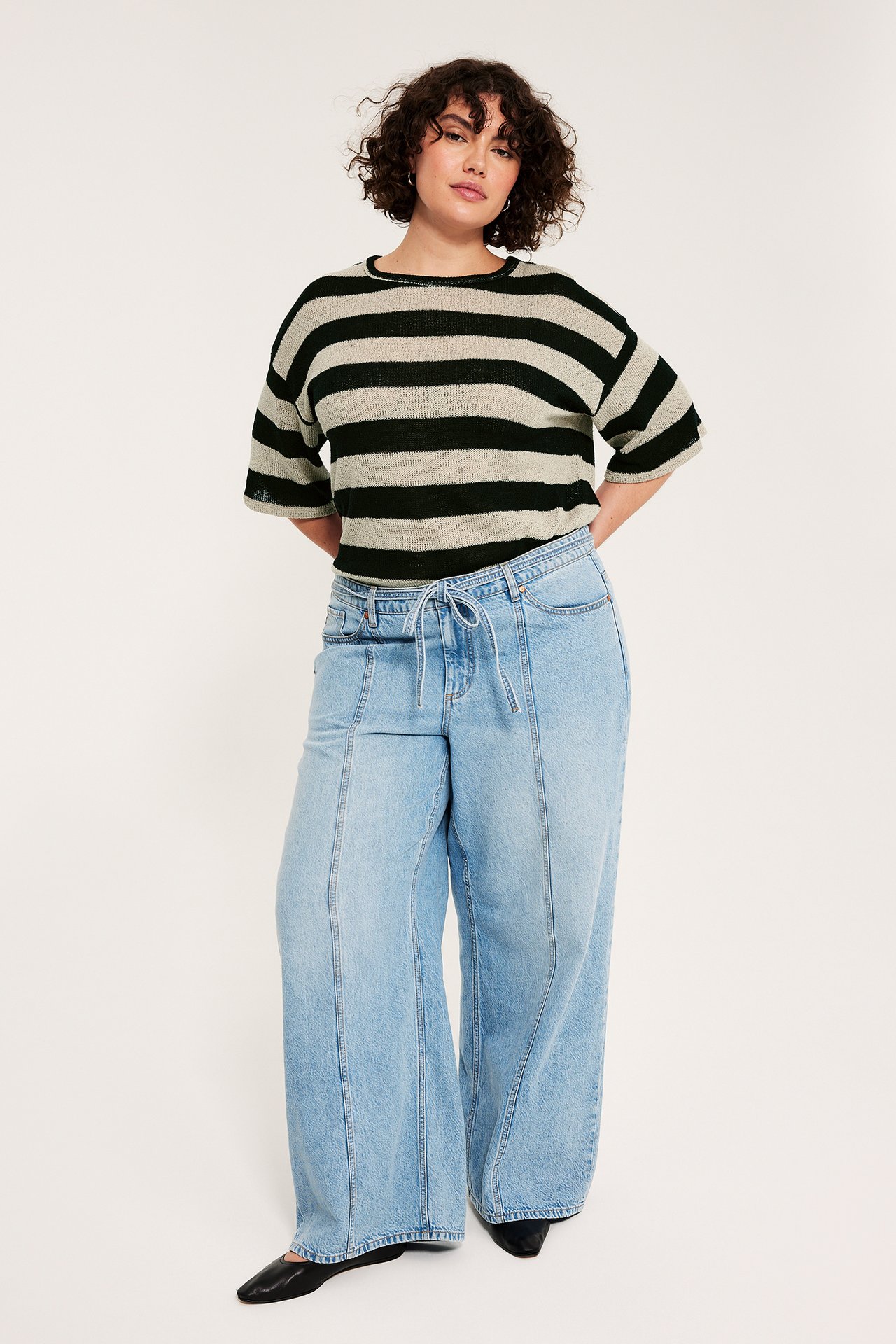 Forfra: kvinne i svart- og lysbeige stripet tynn strikkegenser med korte ermer, lyseblå vide jeans, svarte flate sko.