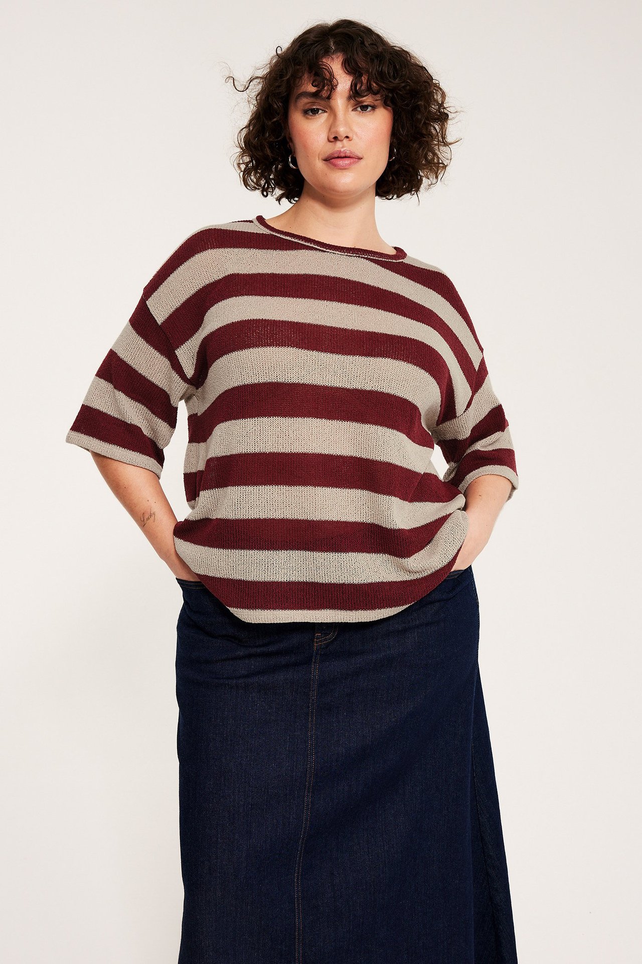 Kobieta w luźnym czerwono-beżowym, pasiastym swetrze z krótkim rękawem i granatowej spódnicy dżinsowej.
