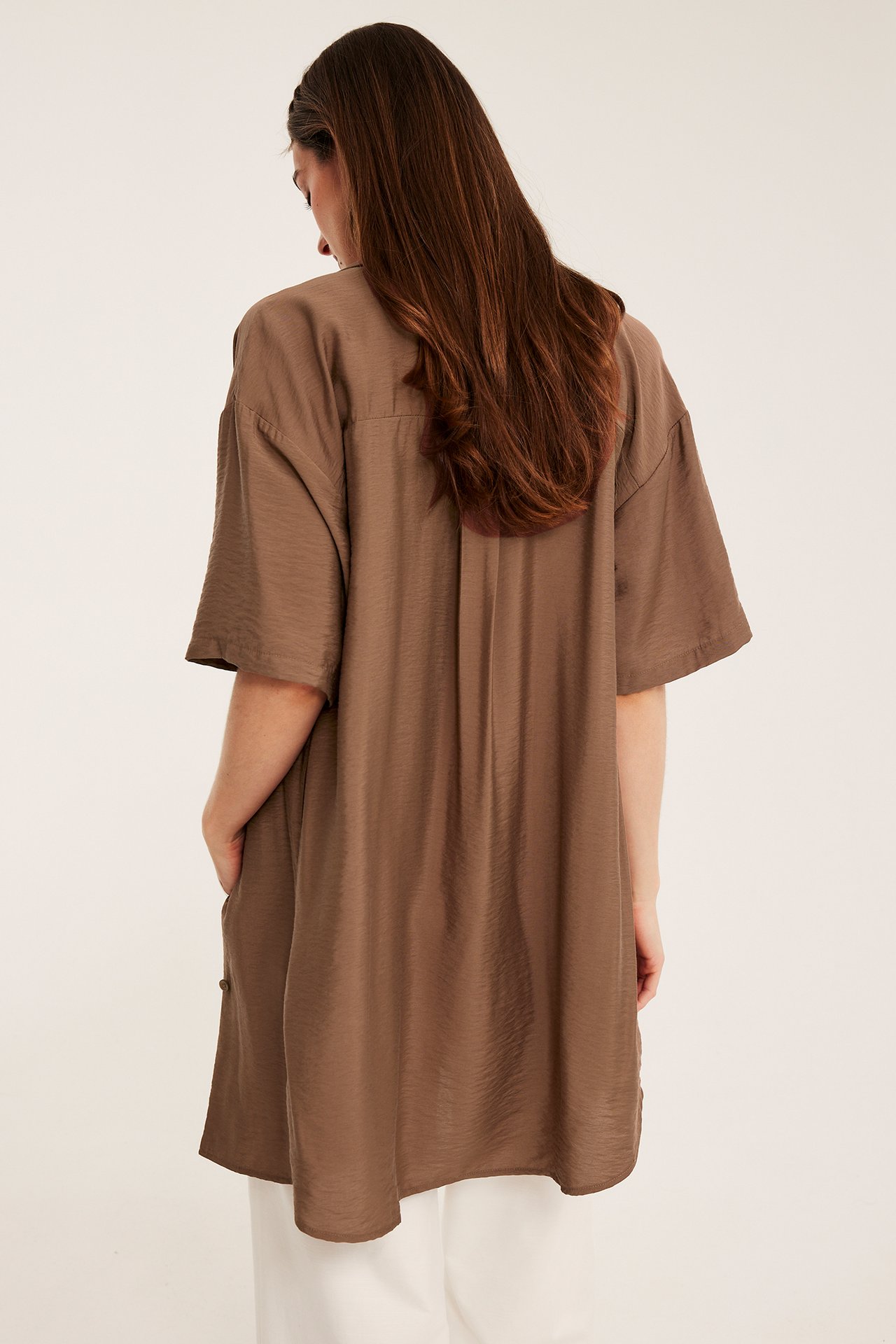 Bakifrån, kvinna i löst sittande, kortärmad brun skjortklänning över vita byxor.