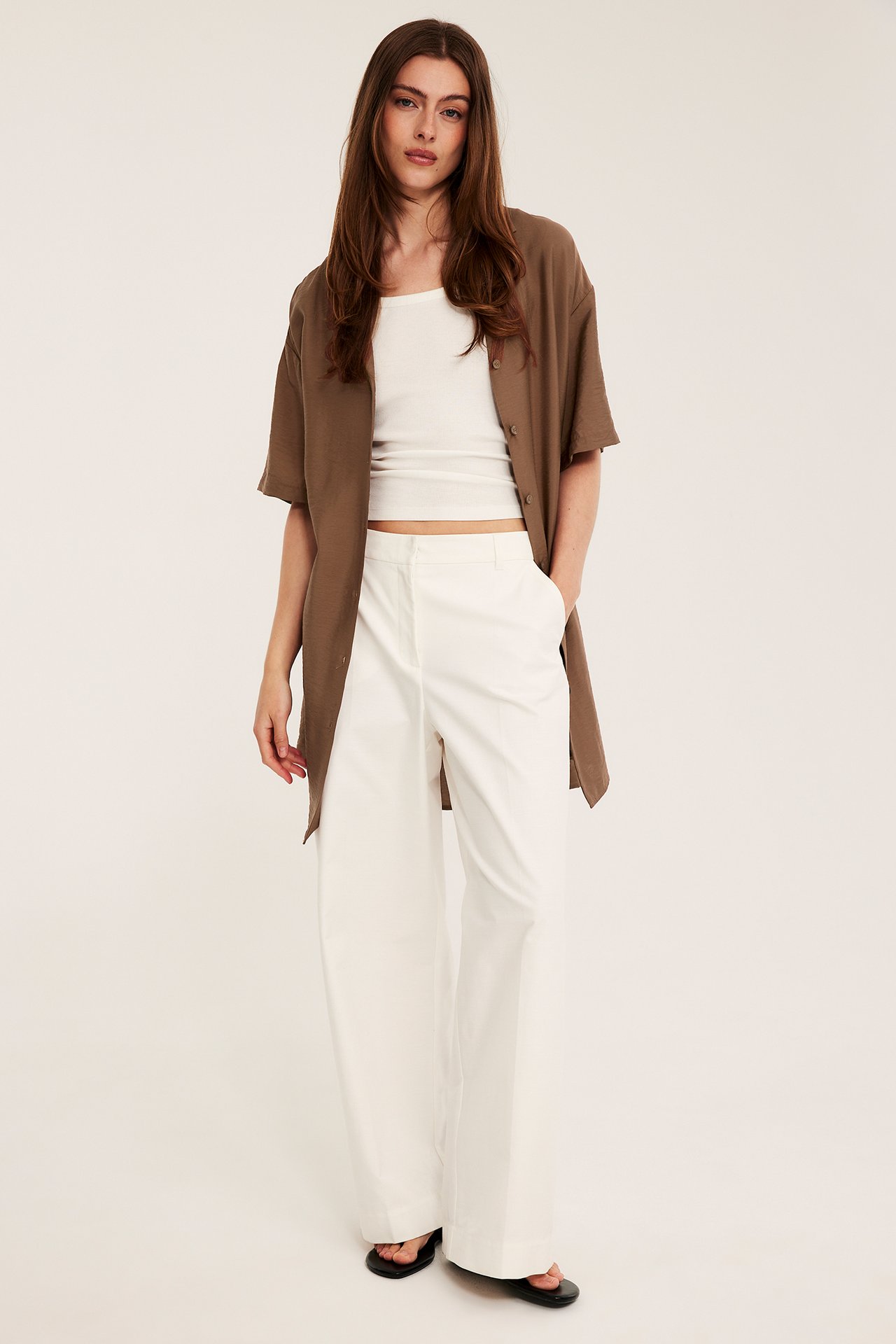 Front view: Kvinna i lös brun kortärmad skjortklänning, öppen över vit linne och vida byxor.