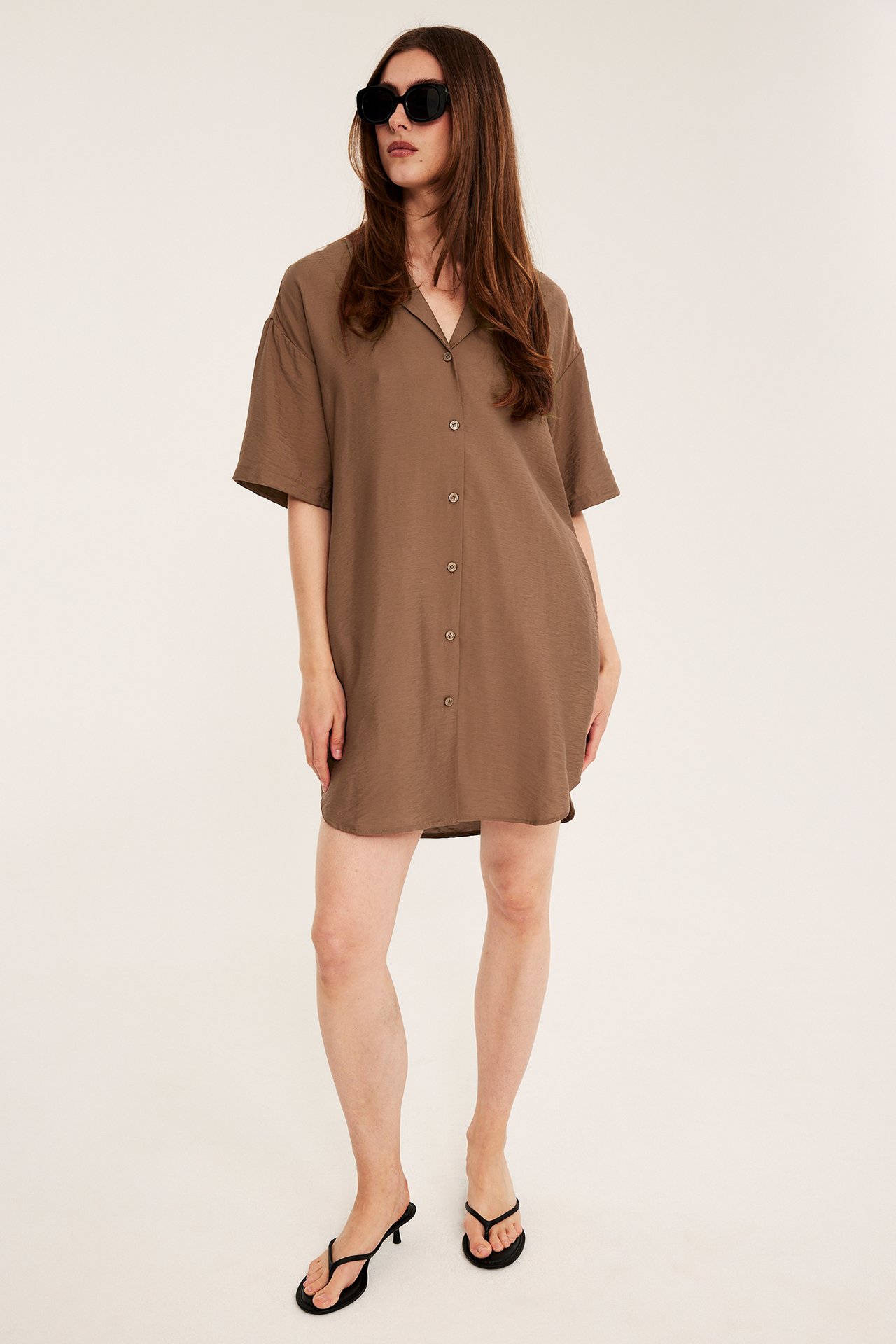 Framifrån, kvinna i brun kortärmad skjortklänning, svarta solglasögon och svarta klackflipflops.