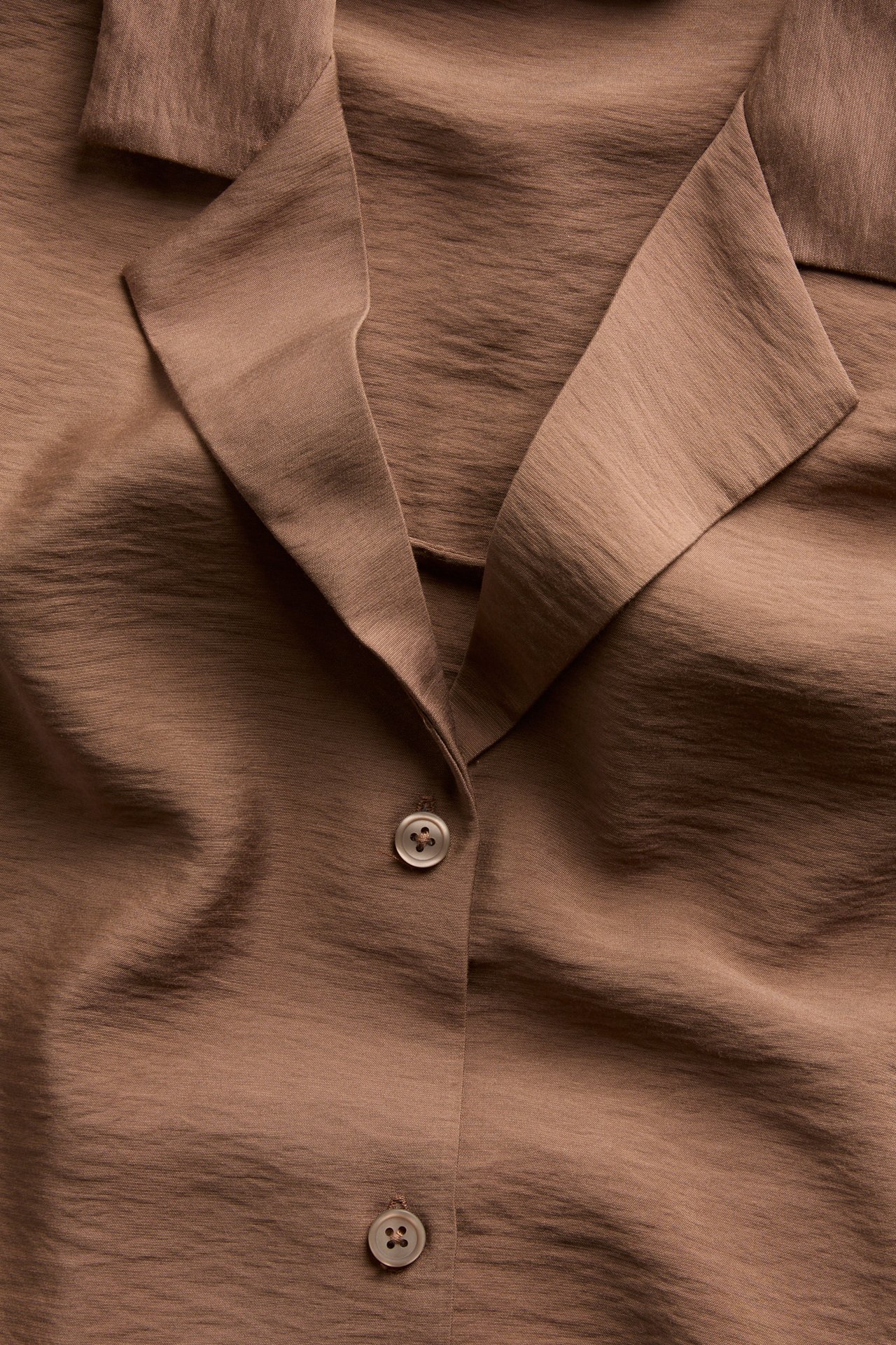 Närbild framifrån på en brun texturerad skjortklänning med öppen krage och två ljusa knappar.