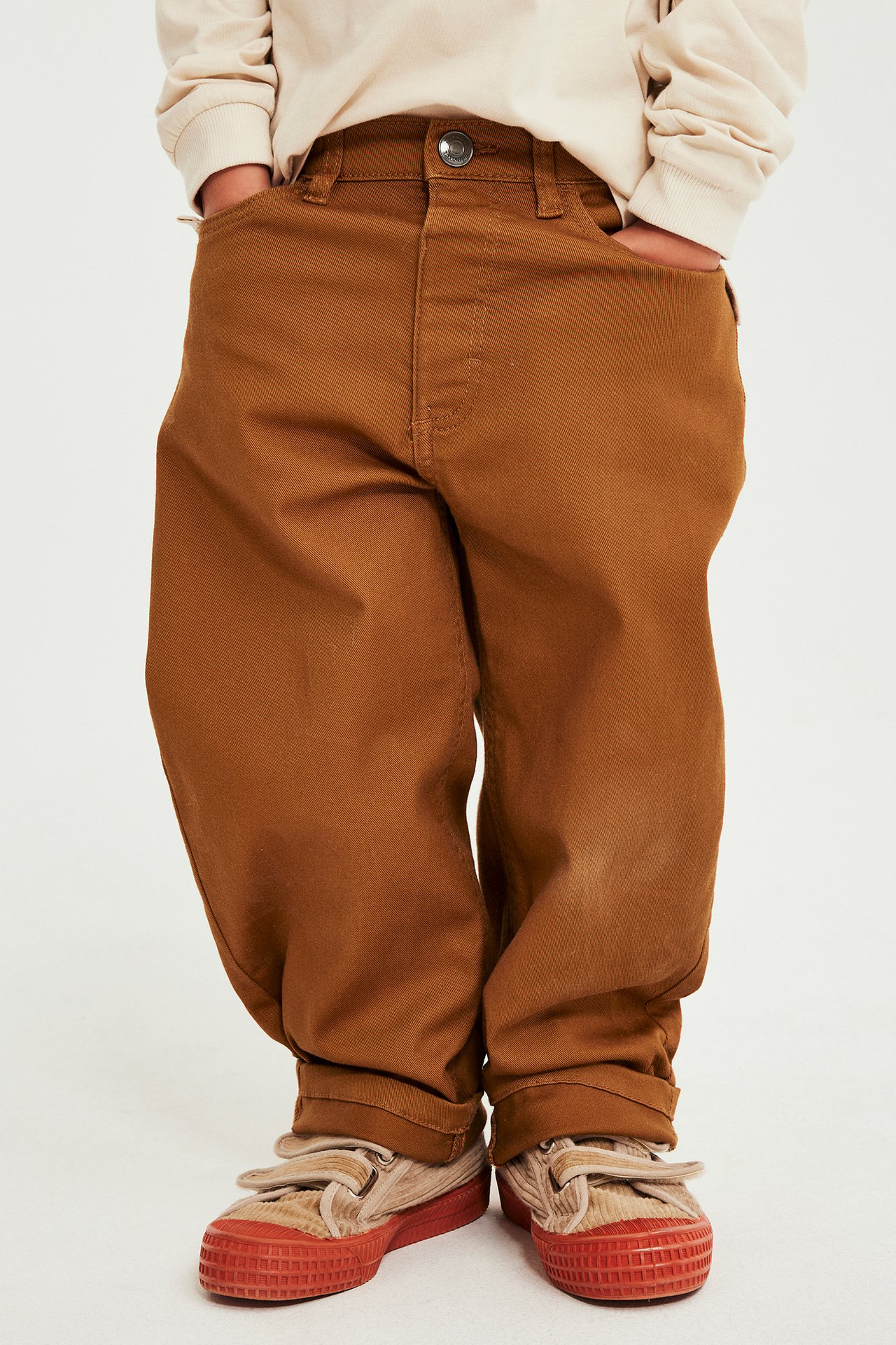 Framifrån, barn i bruna vida bomullsbyxor, beige långärmad topp och beige/röda kardborresneakers.