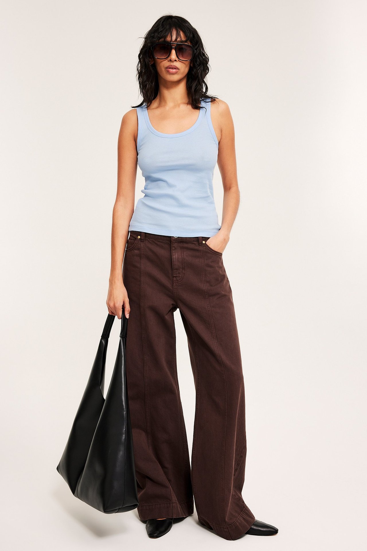 Etukuva: Nainen yllään vaaleansininen ribbitoppi, tummanruskeat leveät housut, aurinkolasit, pitää mustaa kassia.