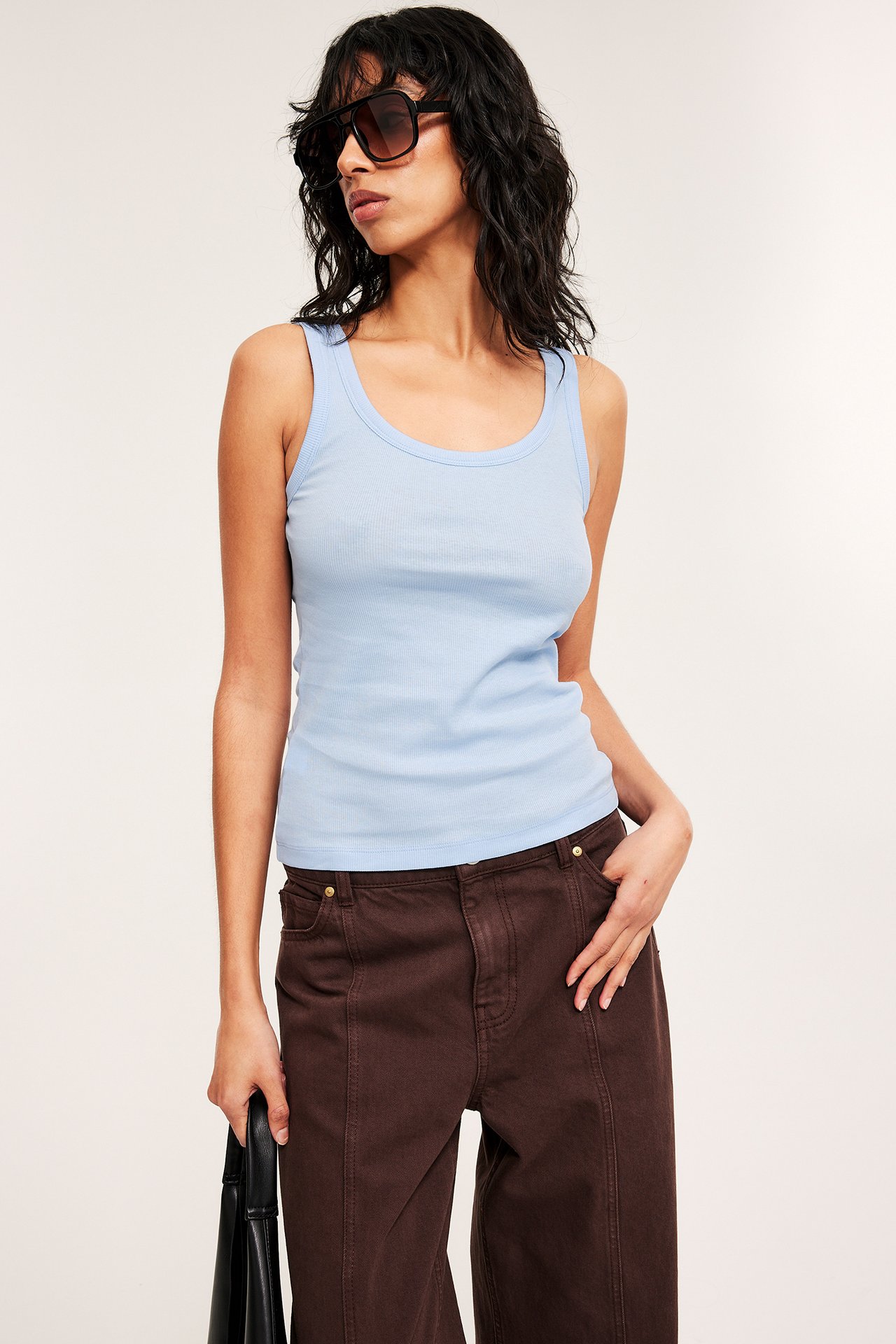 Edestä: nainen yllään vaaleansininen ribbitoppi, ruskeat leveälahkeiset housut, mustat aurinkolasit, pitää mustaa laukkua.