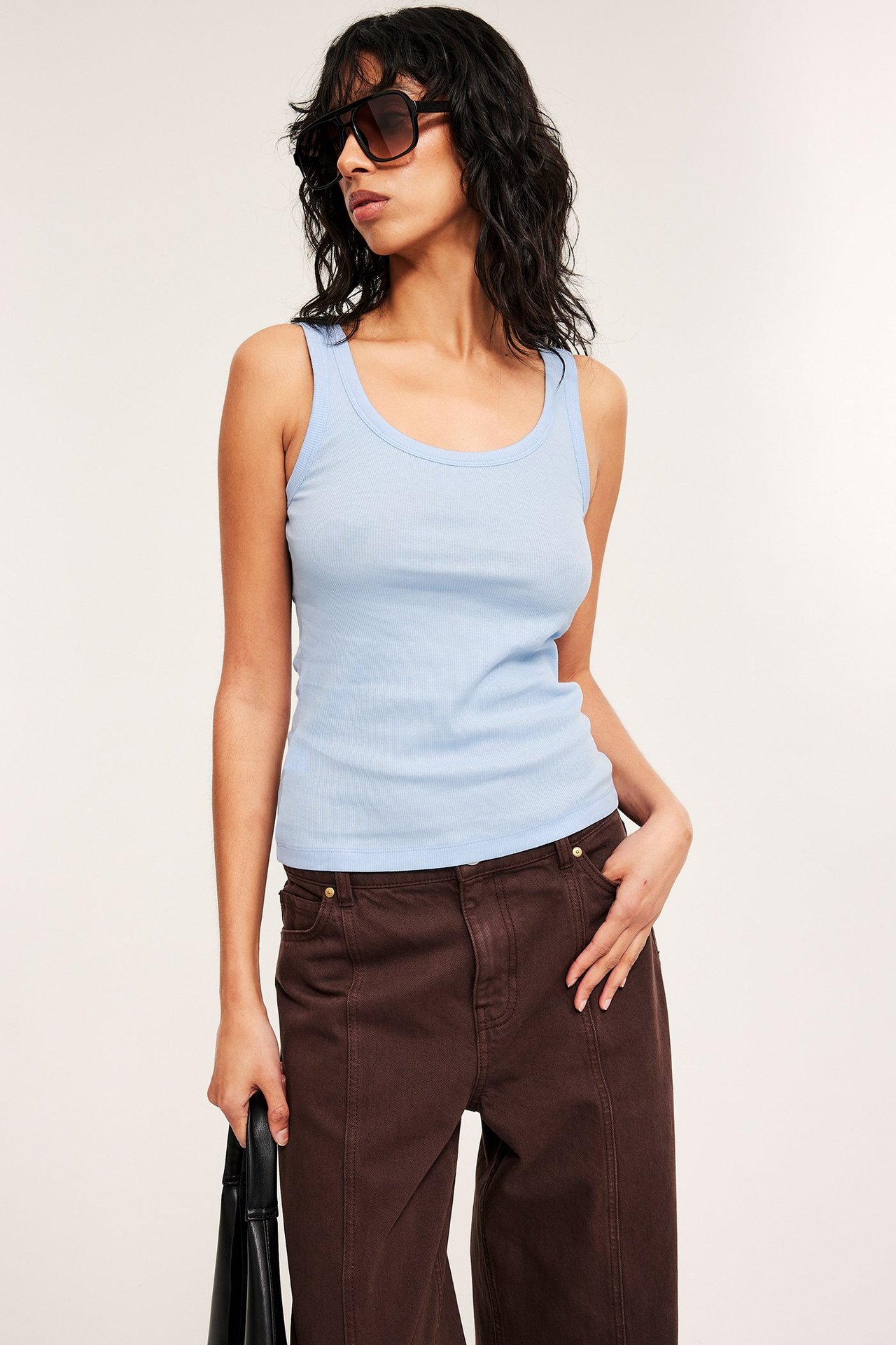 Edestä: nainen yllään vaaleansininen ribbitoppi, ruskeat leveälahkeiset housut, mustat aurinkolasit, pitää mustaa laukkua.