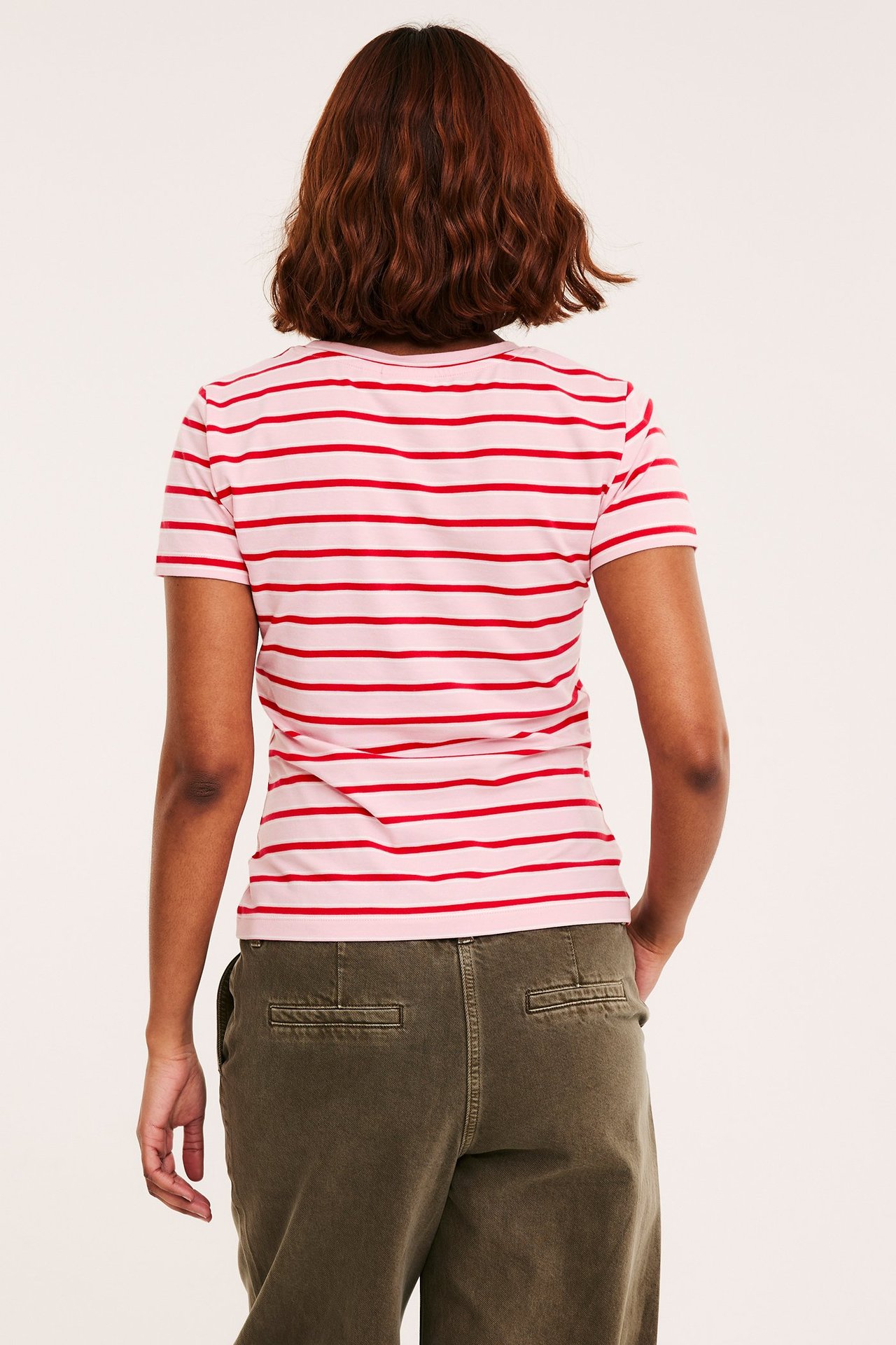 Naisen takakuva yllään istuva vaaleanpunainen ja punaraitainen t-paita sekä oliivinvihreät housut.