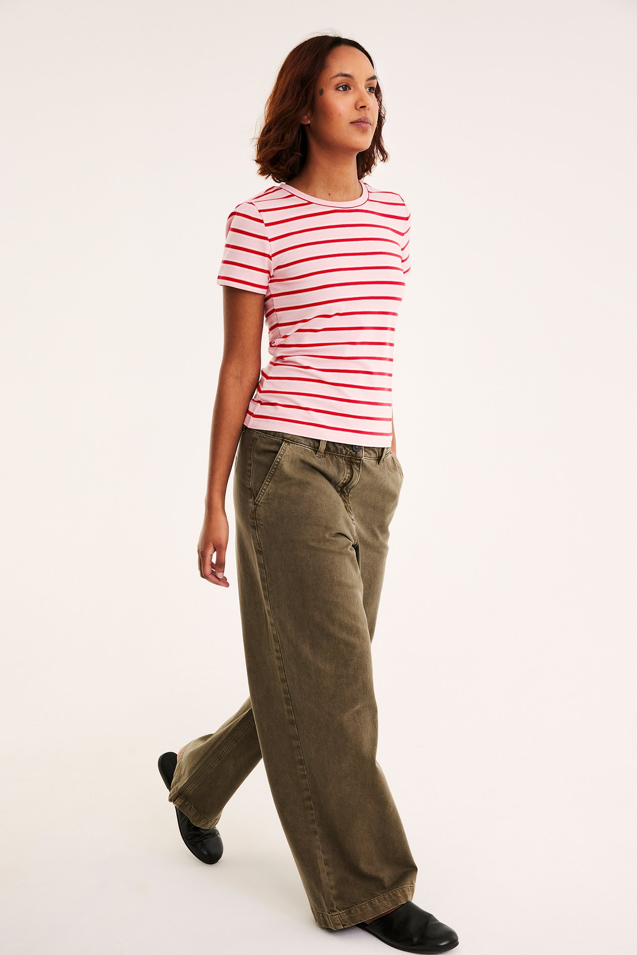Naisen sivu-etunäkymä, yllään istuva vaaleanpunainen-punaraitainen t-paita, oliivinvihreät leveälahkeiset housut, mustat kengät.