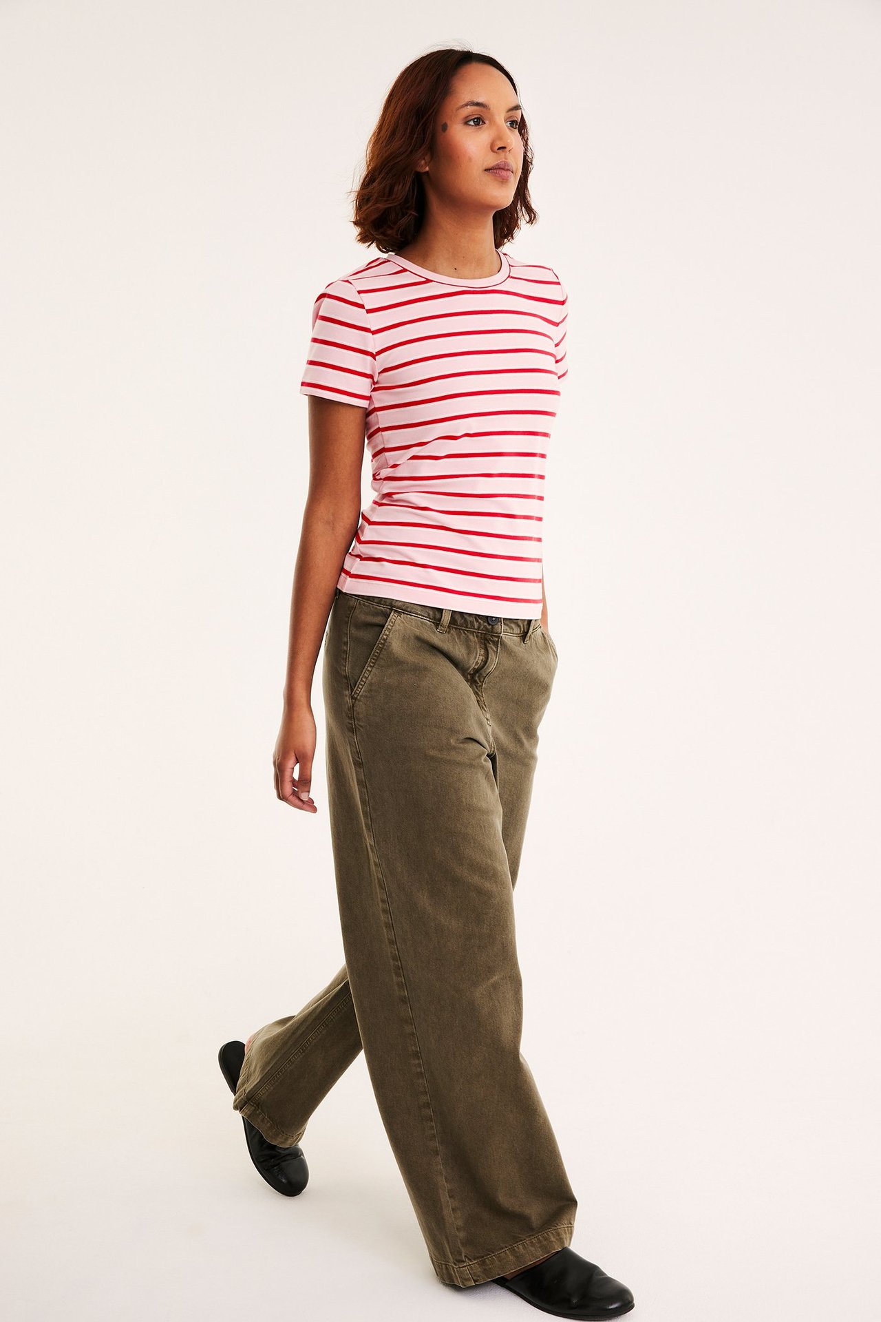 Sidefra-visning av en kvinne i figurnær rosa-rødstripet t-skjorte, olivengrønne vide bukser og svarte flate sko.