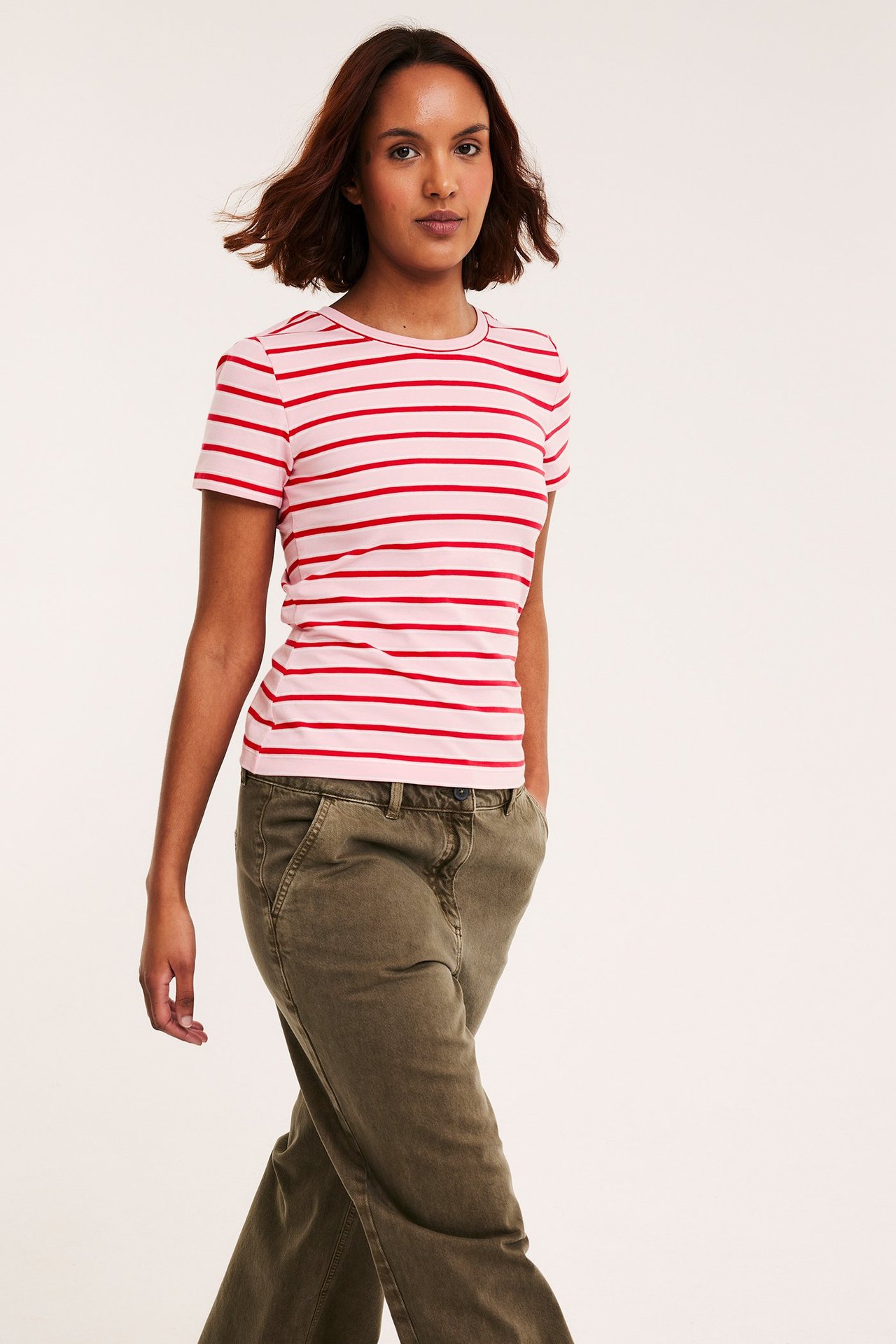 Forfra: Kvinne i ettersittende rosa og rødstripet t-skjorte med korte ermer og olivengrønne bukser.