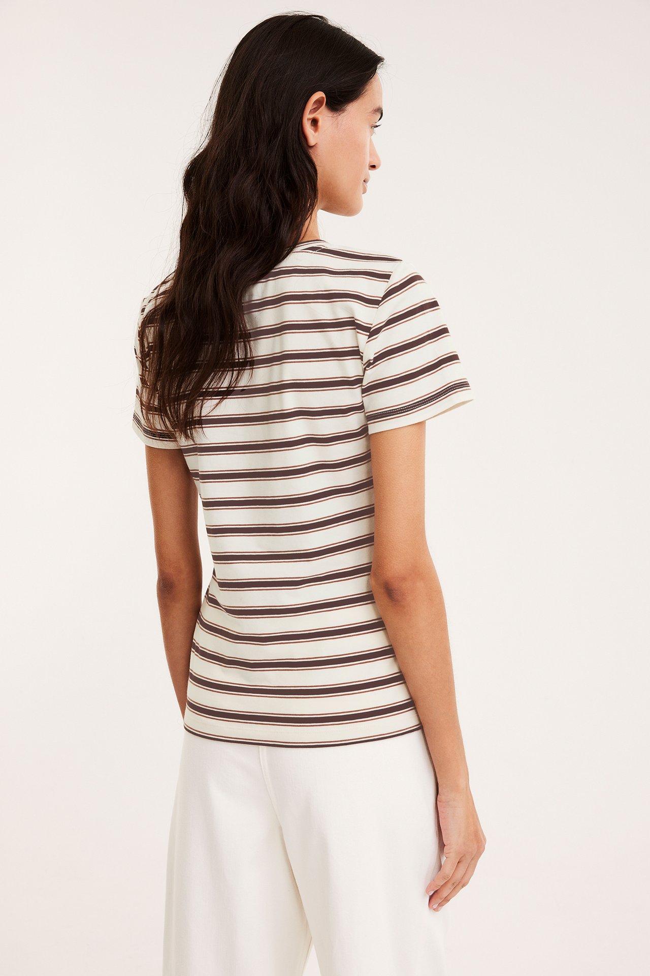 Widok z tyłu: kobieta w dopasowanym biało-brązowym T-shircie w paski i jasnych szerokich spodniach.