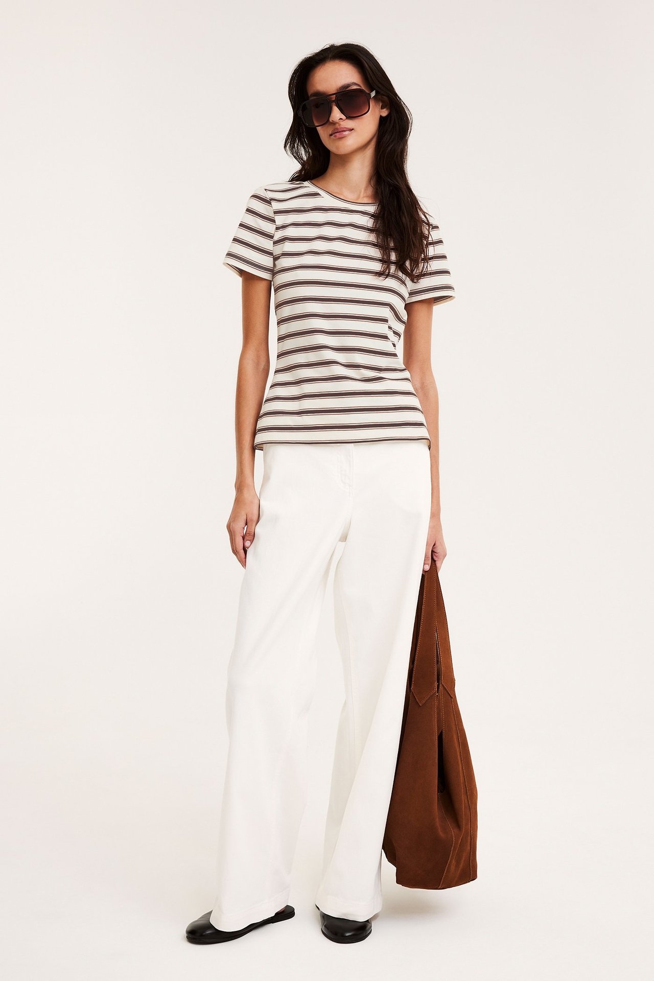 Forfra, kvinne i en figurnær stripete t-skjorte i offwhite, hvite vide bukser, solbriller og holder en brun veske.