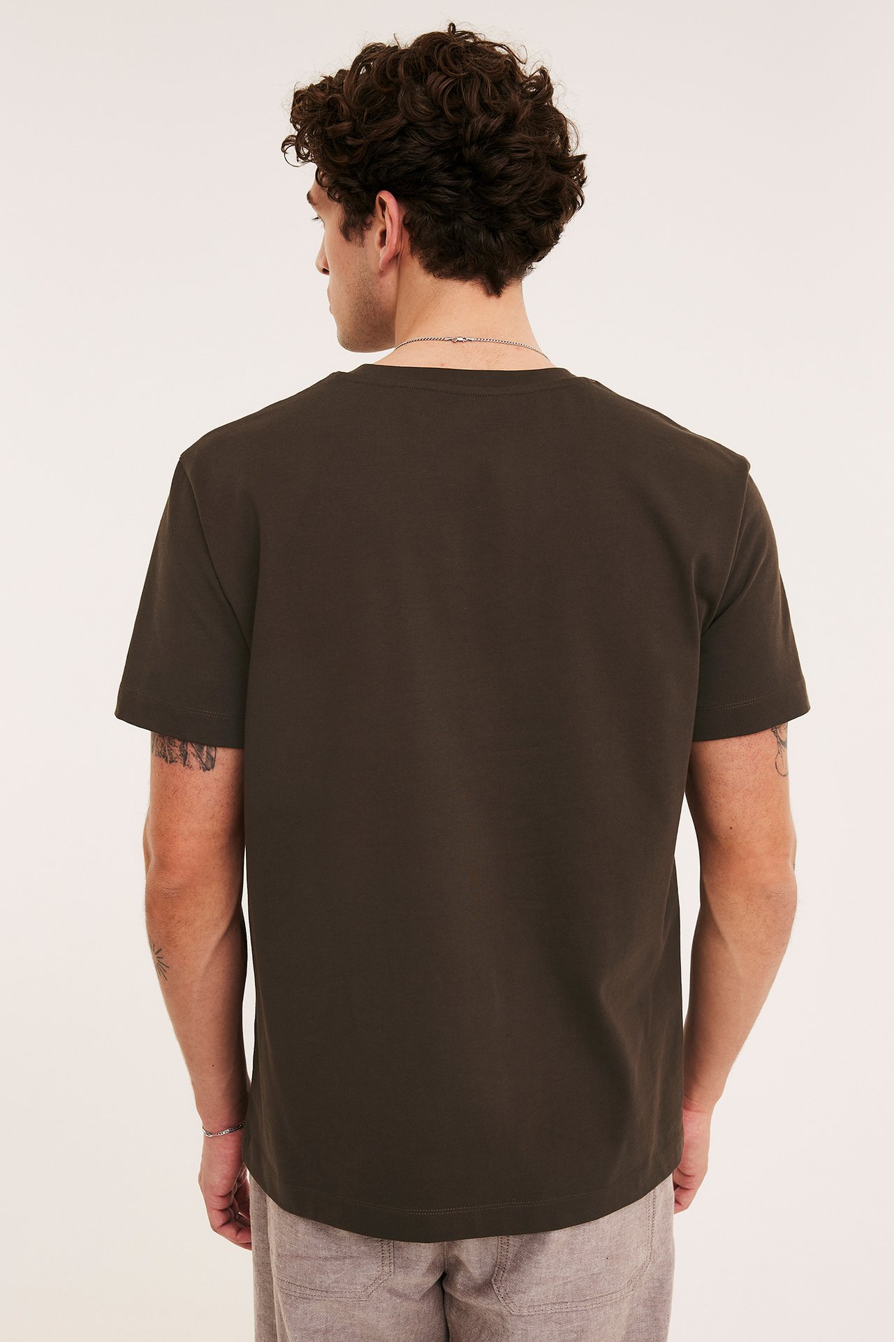 Bakfra, mann i en brun løstsittende bomulls-T-skjorte, beigefargede shorts og et sølvkjede.