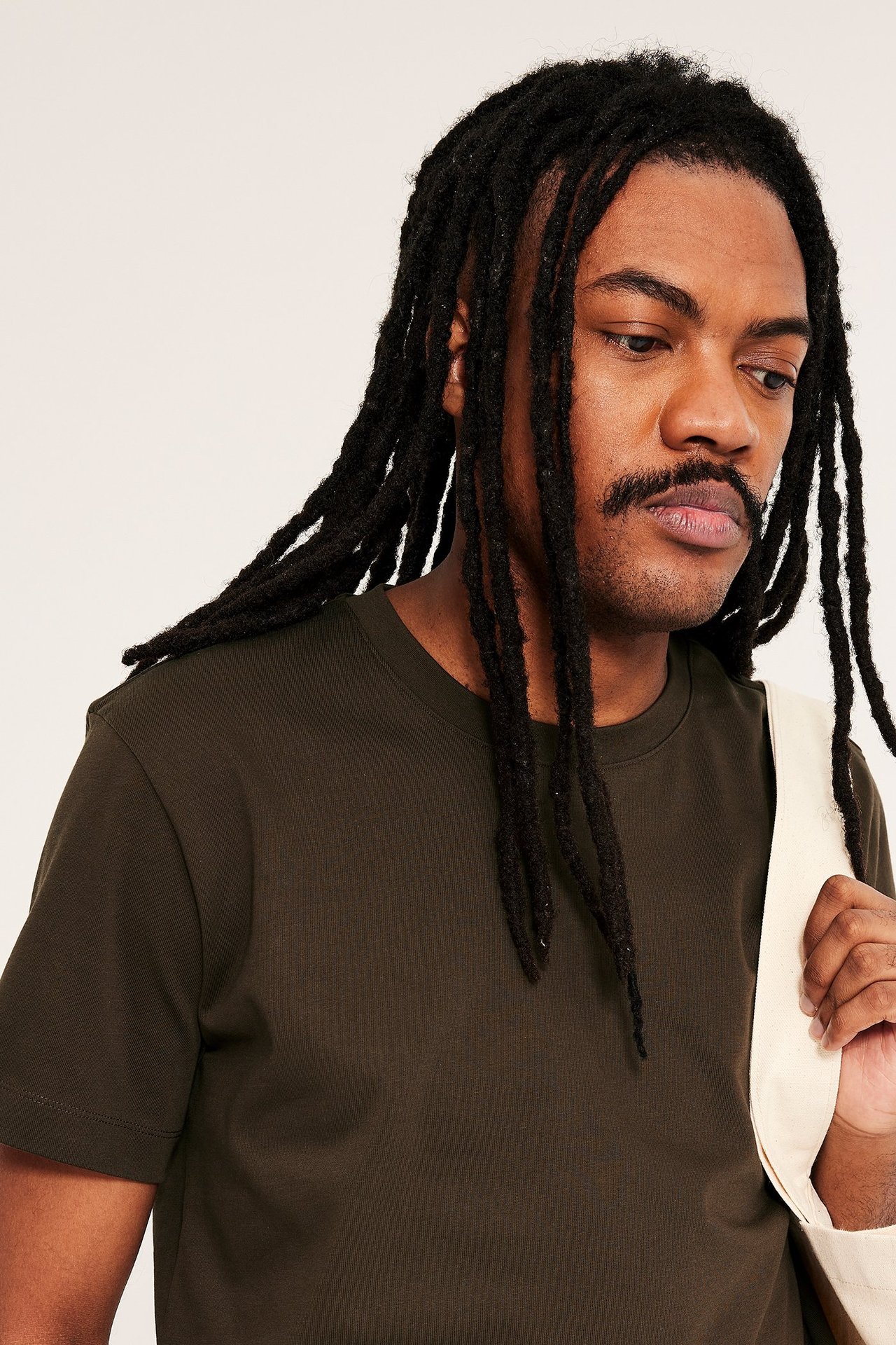 Närbild, sidovy av man i mörkbrun lös passform O-ringad bomulls-t-shirt, håller en beige tygkasse.