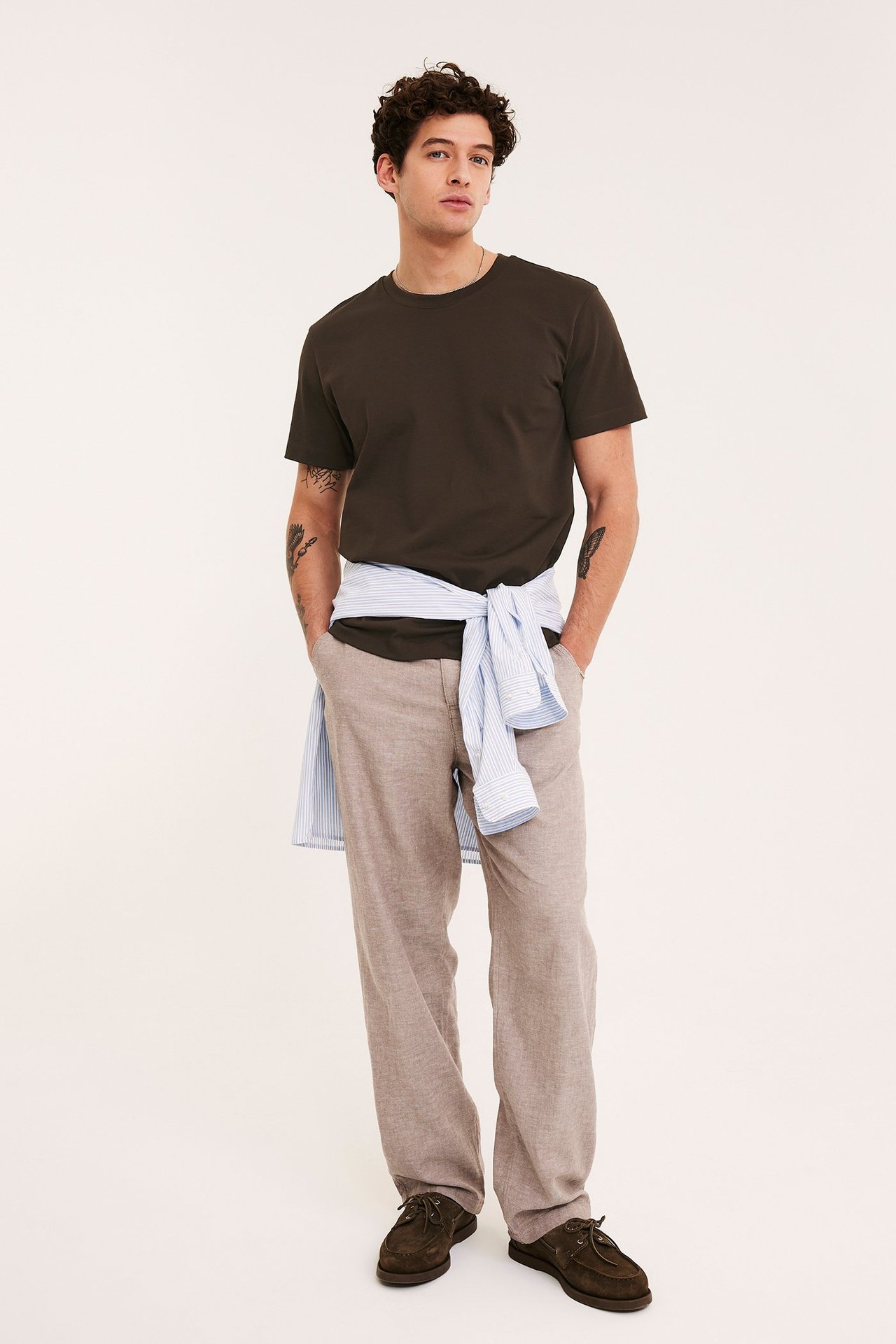 Forfra: Mann i brun løs t-skjorte, beige bukser, mørke sko. Stripet skjorte knyttet rundt midjen.