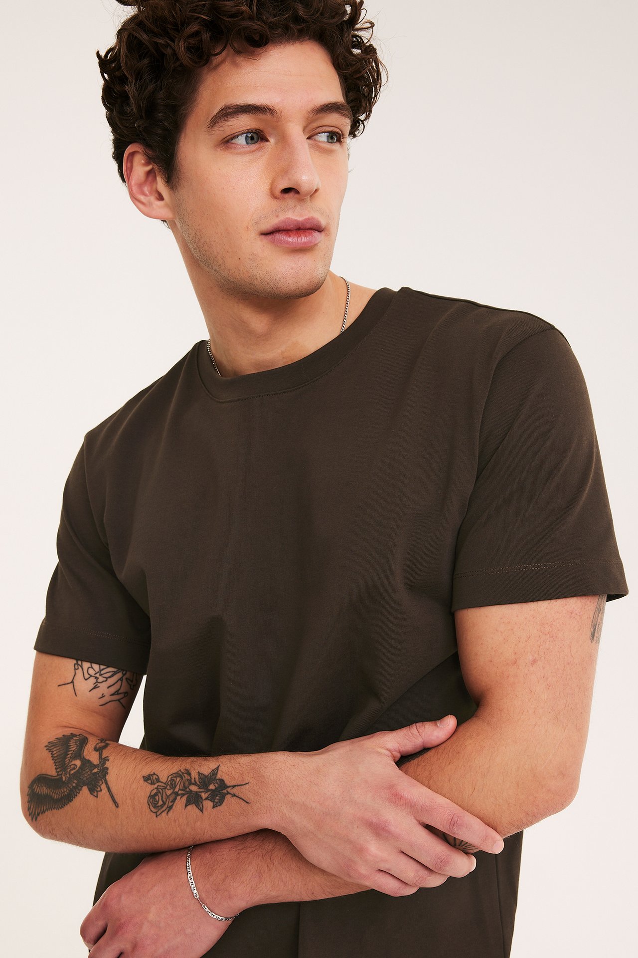 Nærbilde forfra av en mann iført en løstsittende brun O-hals bomulls-T-skjorte, et sølvkjede og et sølvarmbånd.