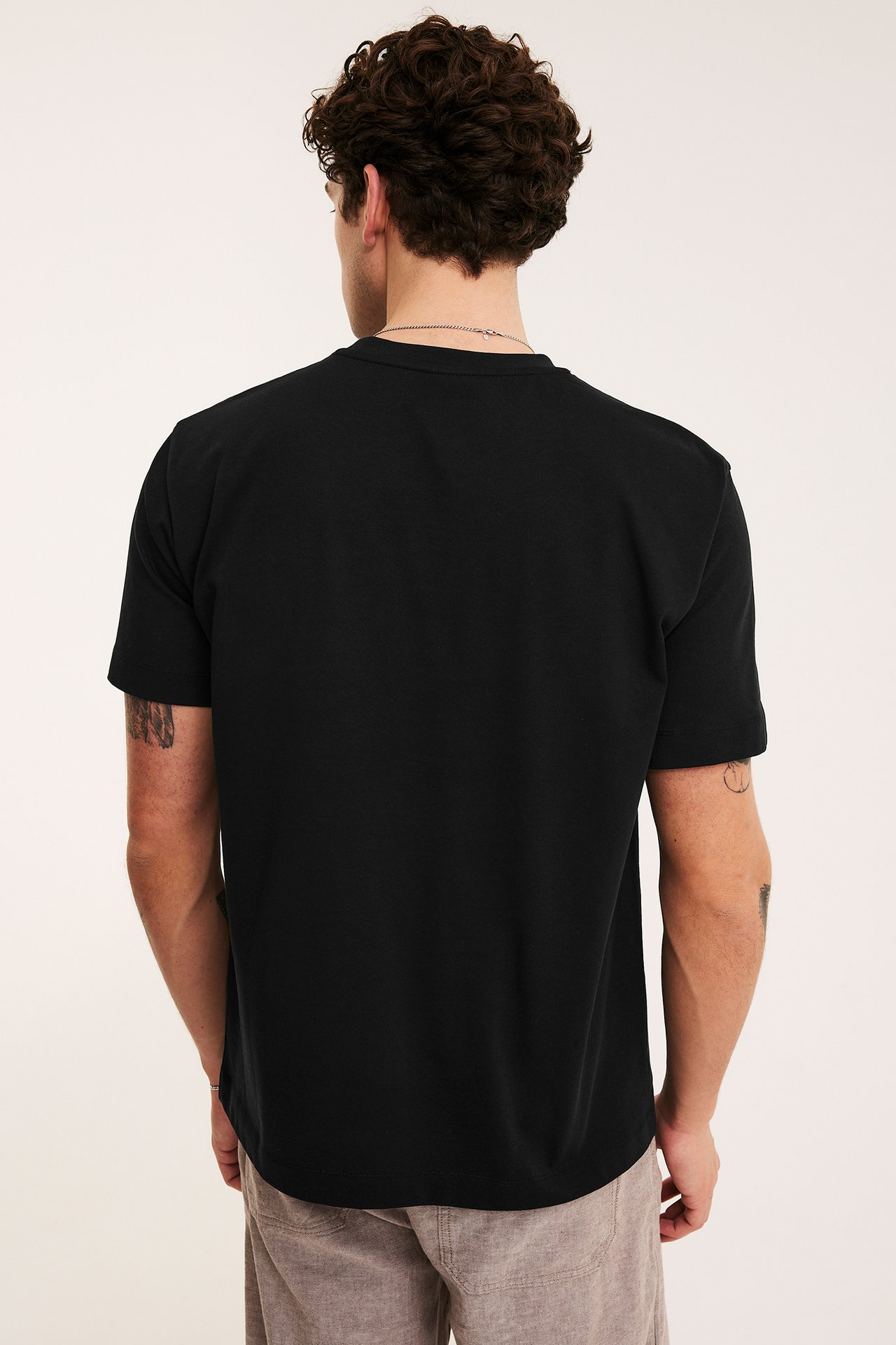 Widok z tyłu: mężczyzna w czarnym, luźnym t-shircie O-neck, beżowych spodniach, srebrnym łańcuszku.