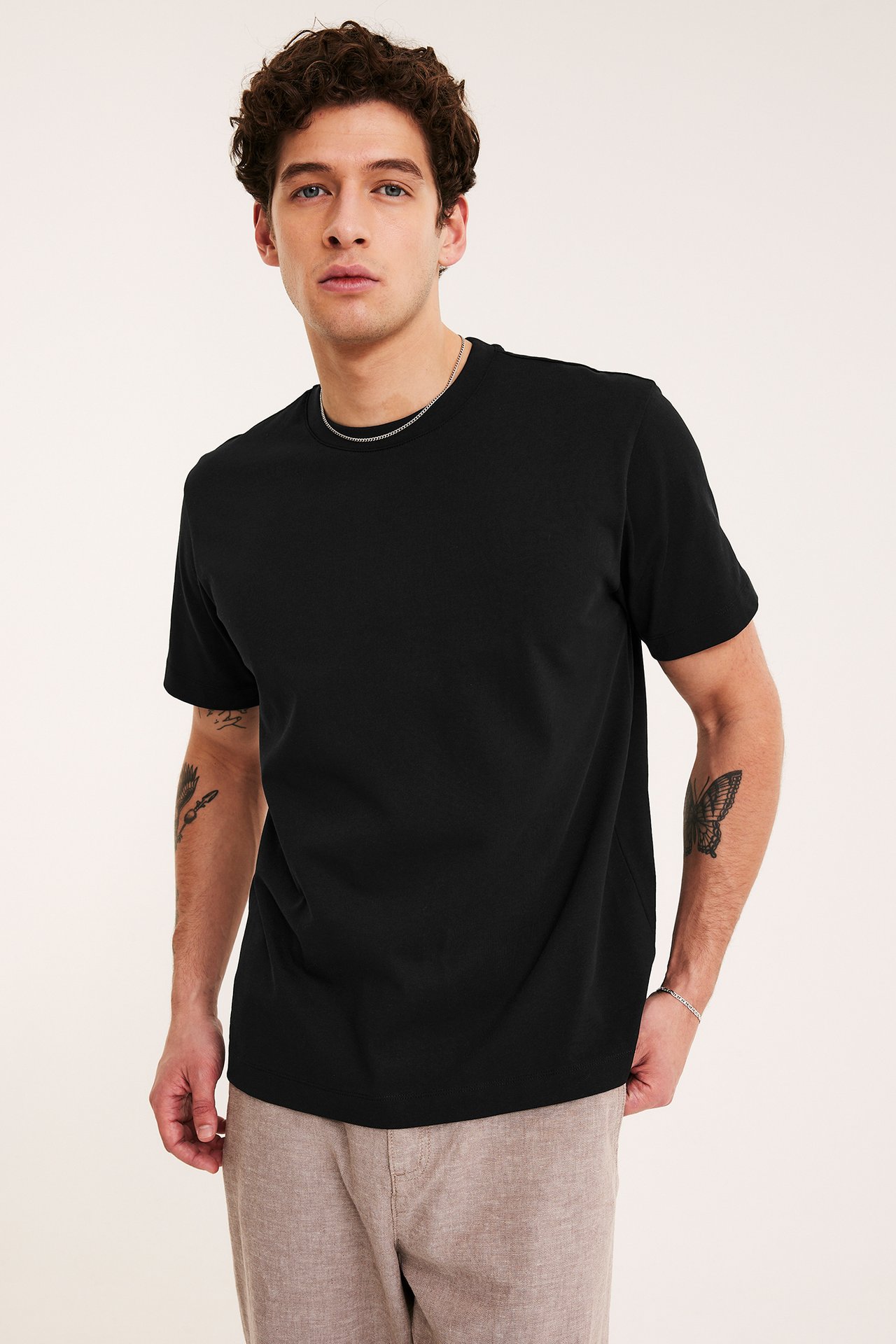 Widok z przodu mężczyzny w czarnej luźnej koszulce z okrągłym dekoltem, beżowych spodniach i srebrnym naszyjniku.