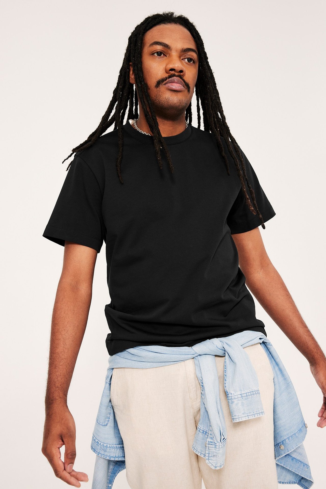 Man i svart lös T-shirt, beiga byxor och ljusblå jeansskjorta knuten i midjan.