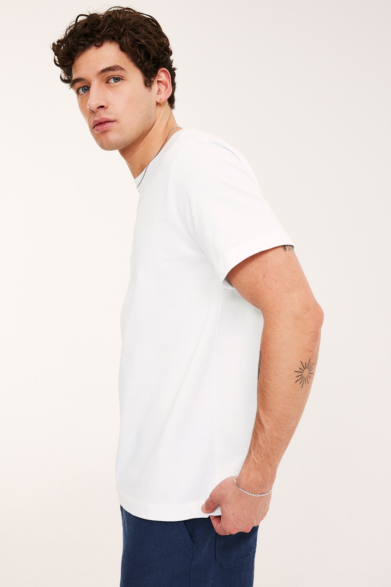 Mężczyzna, widok z boku: biały, luźny, bawełniany T-shirt z okrągłym dekoltem, granatowe szorty, srebrny łańcuszek i bransoletka.