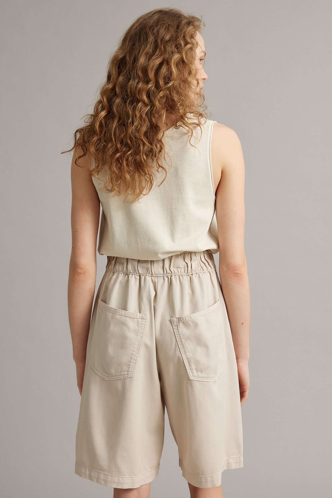 Naisen selkä: vaaleat beiget lyocell-puuvillashortsit joustovyötäröllä ja luonnonvalkoinen hihaton toppi.