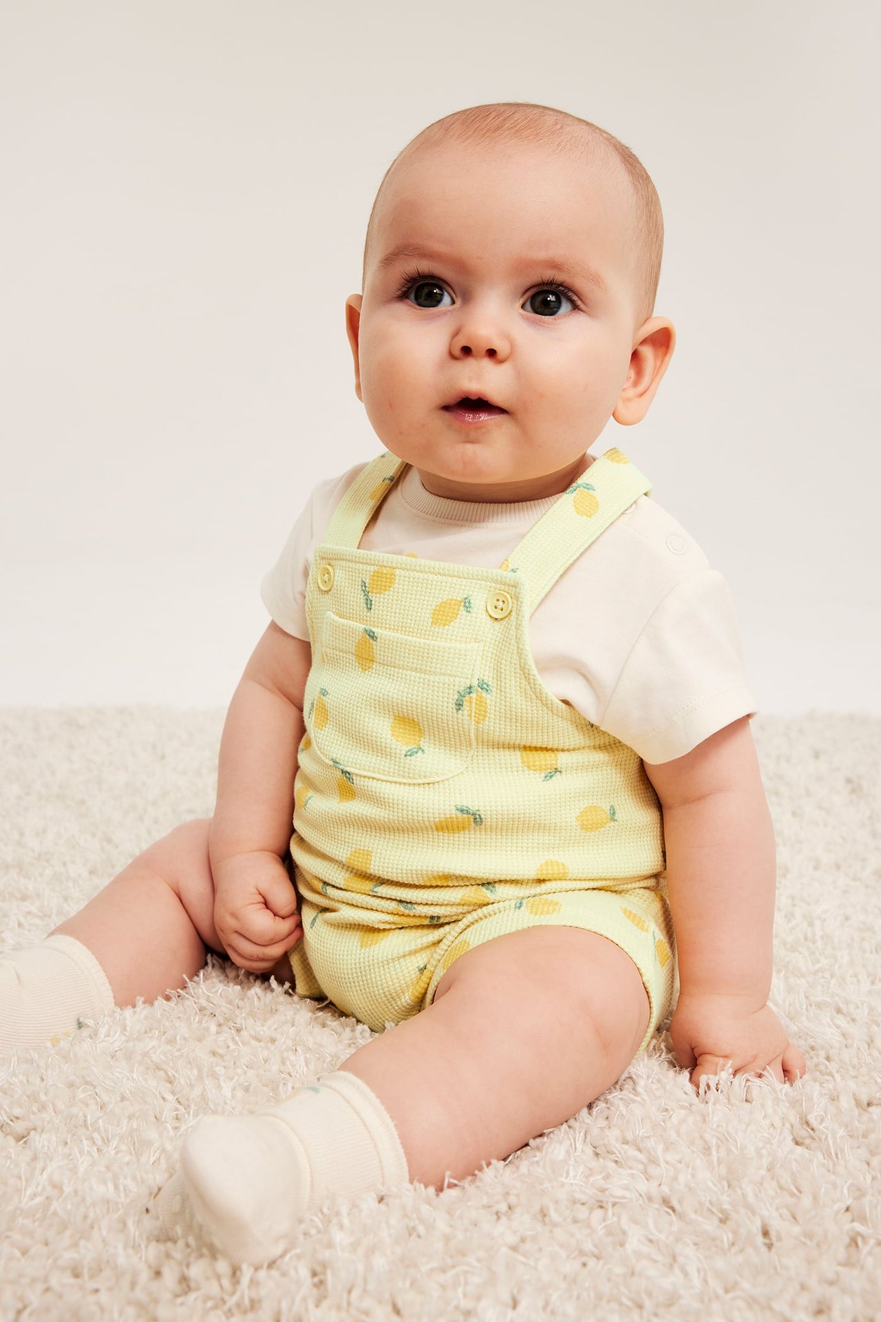 Frontvy: baby i ljusgula våffelhängselbyxor med citronmönster över en ljusbeige t-shirt och strumpor.