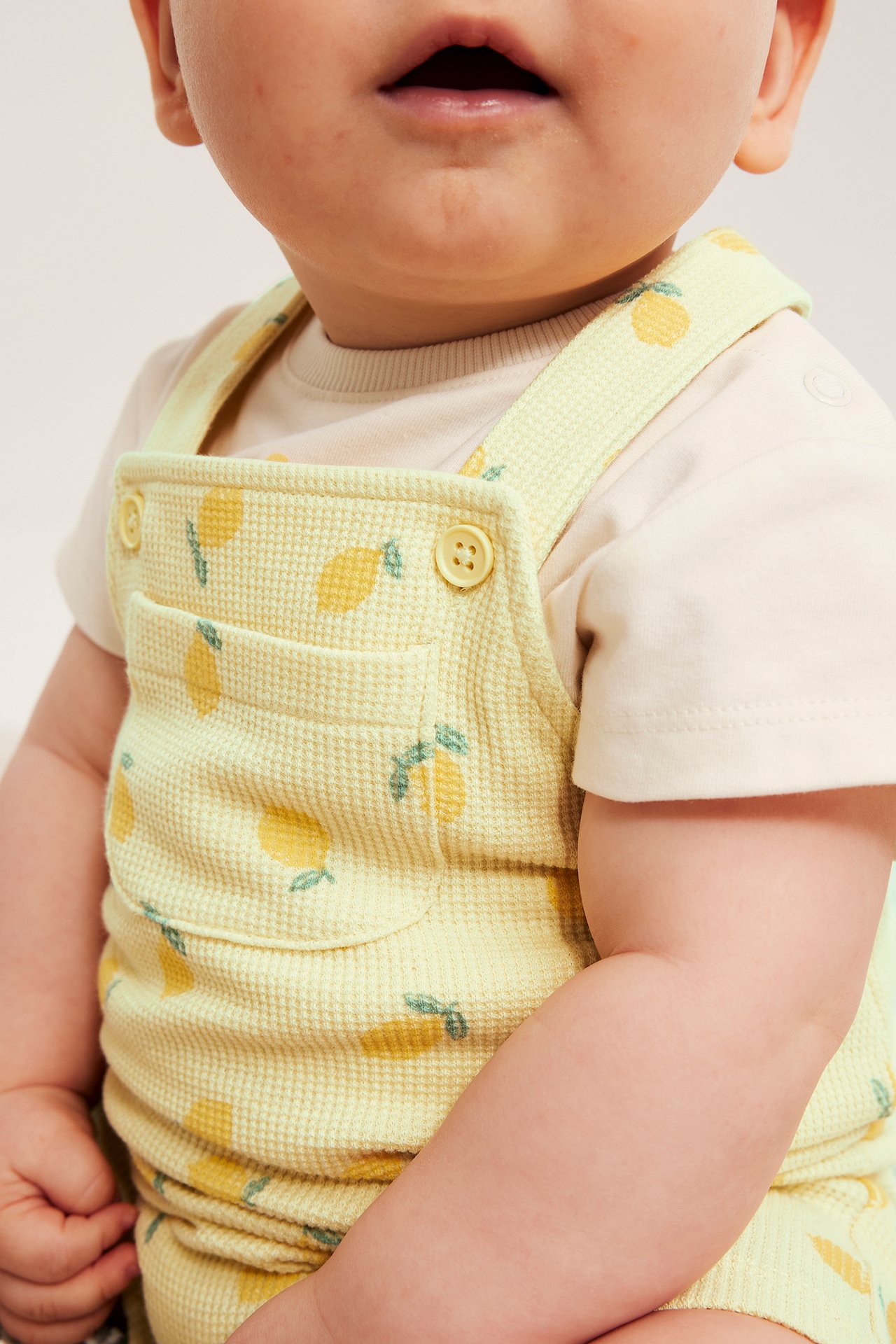 Närbild: baby i ljusgula våffelhängselbyxor med citronmönster och bröstficka, över beige t-shirt.