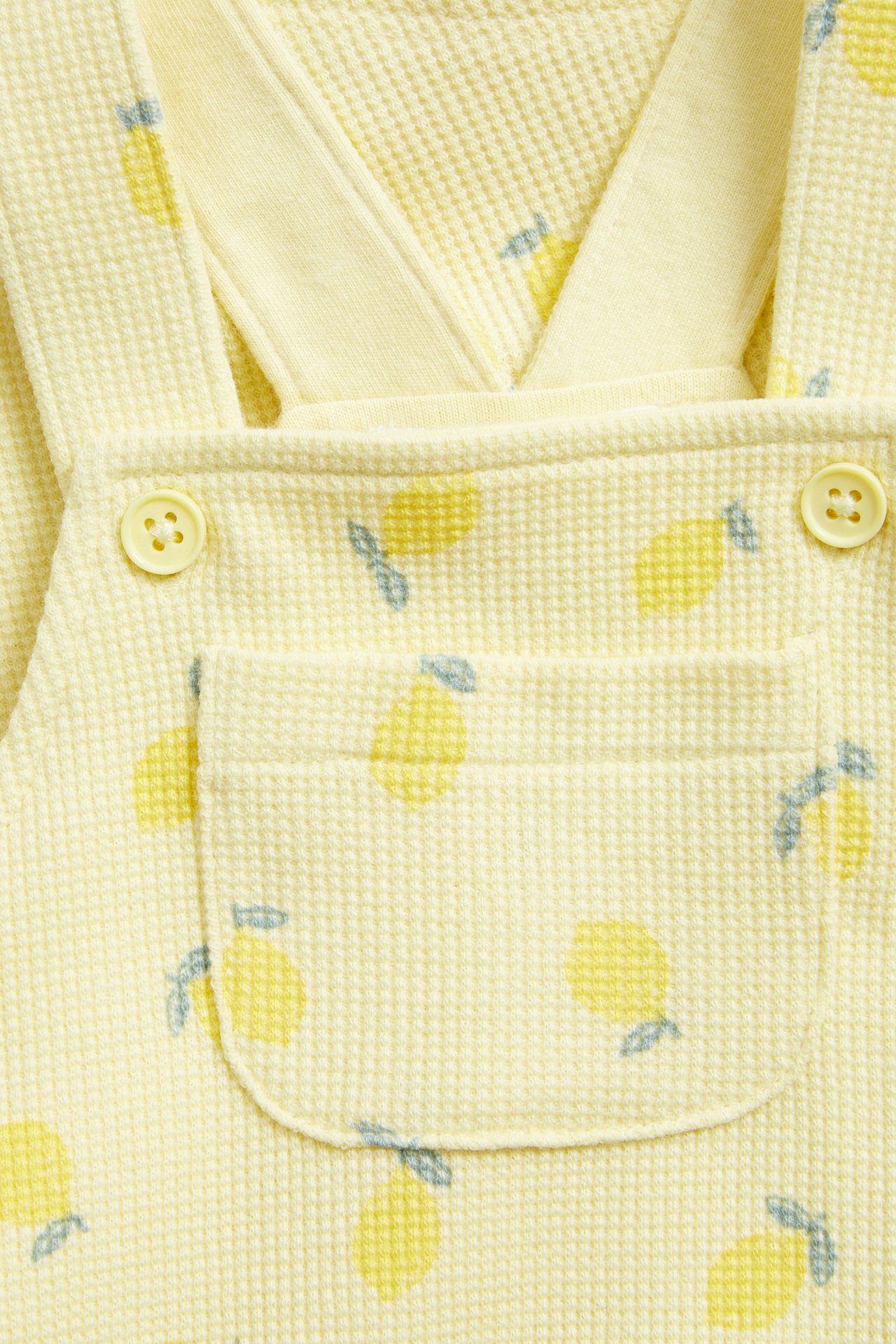 Zbliżenie na jasne, żółte waflowe ogrodniczki niemowlęce w cytrynowy wzór, z guzikami i kieszenią.