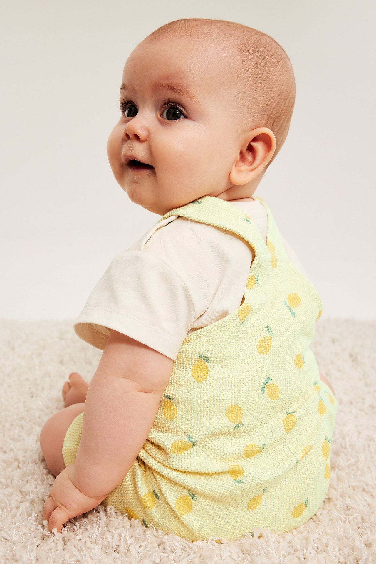 Sidovy av en baby i ljusgul våfflad hängselbyxa med citronmönster över en krämfärgad t-shirt.