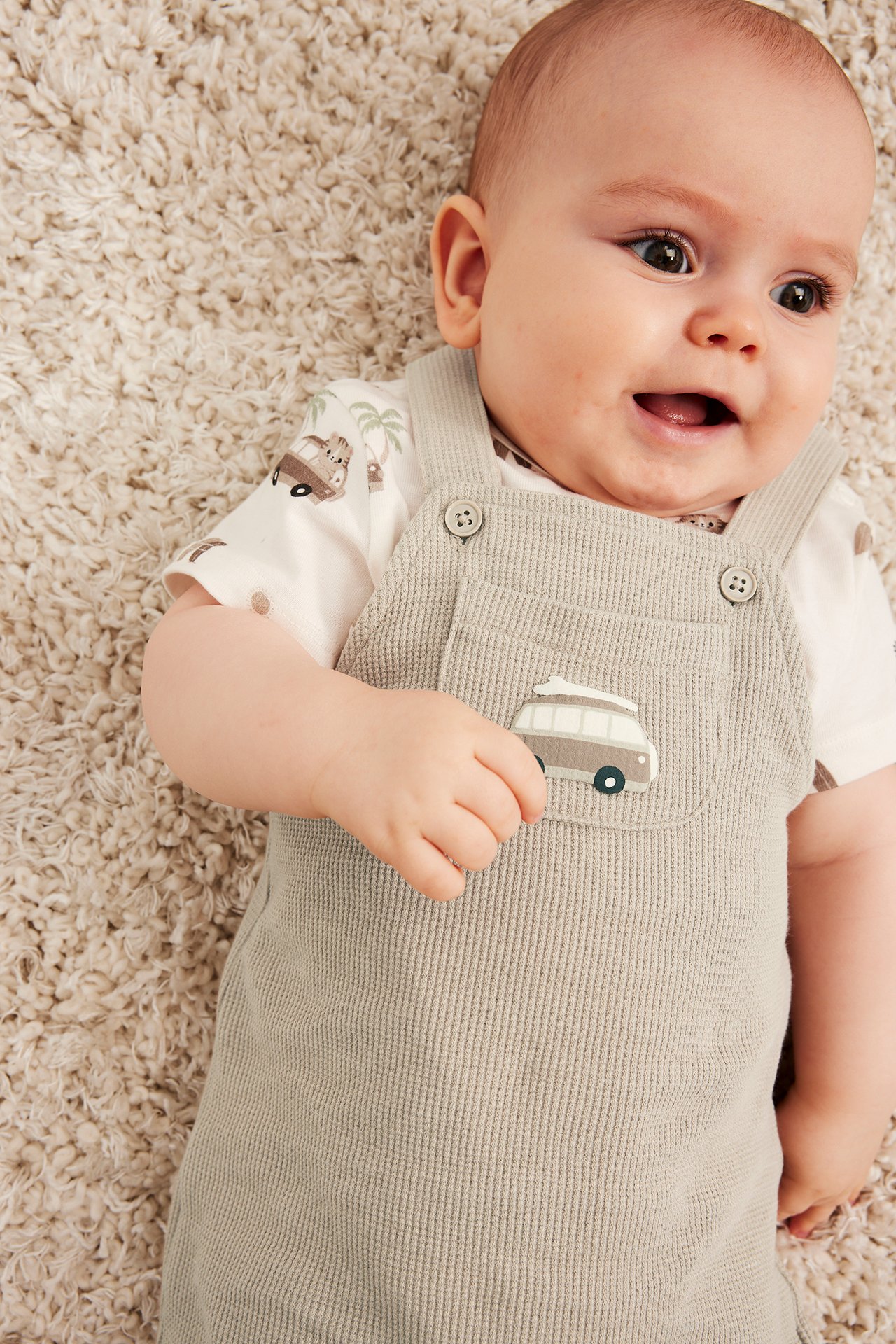 Nærbilde av baby i beige vaffelstrikkede snekkerbukser med bobilapplikasjon og hvit safari-mønstret body.