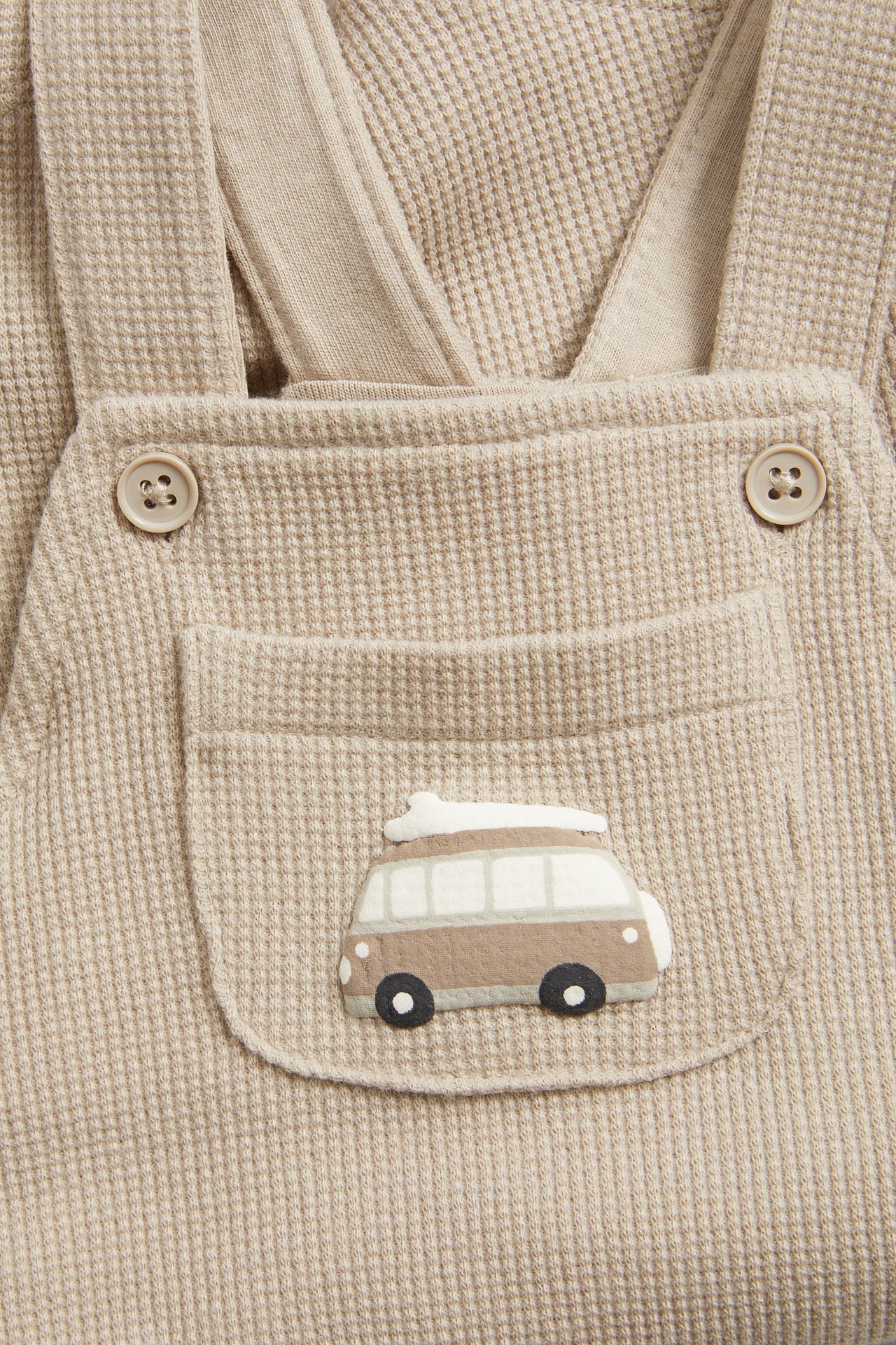 Z przodu: beżowe waflowe ogrodniczki niemowlęce z guzikami i kieszenią z aplikacją kampera.
